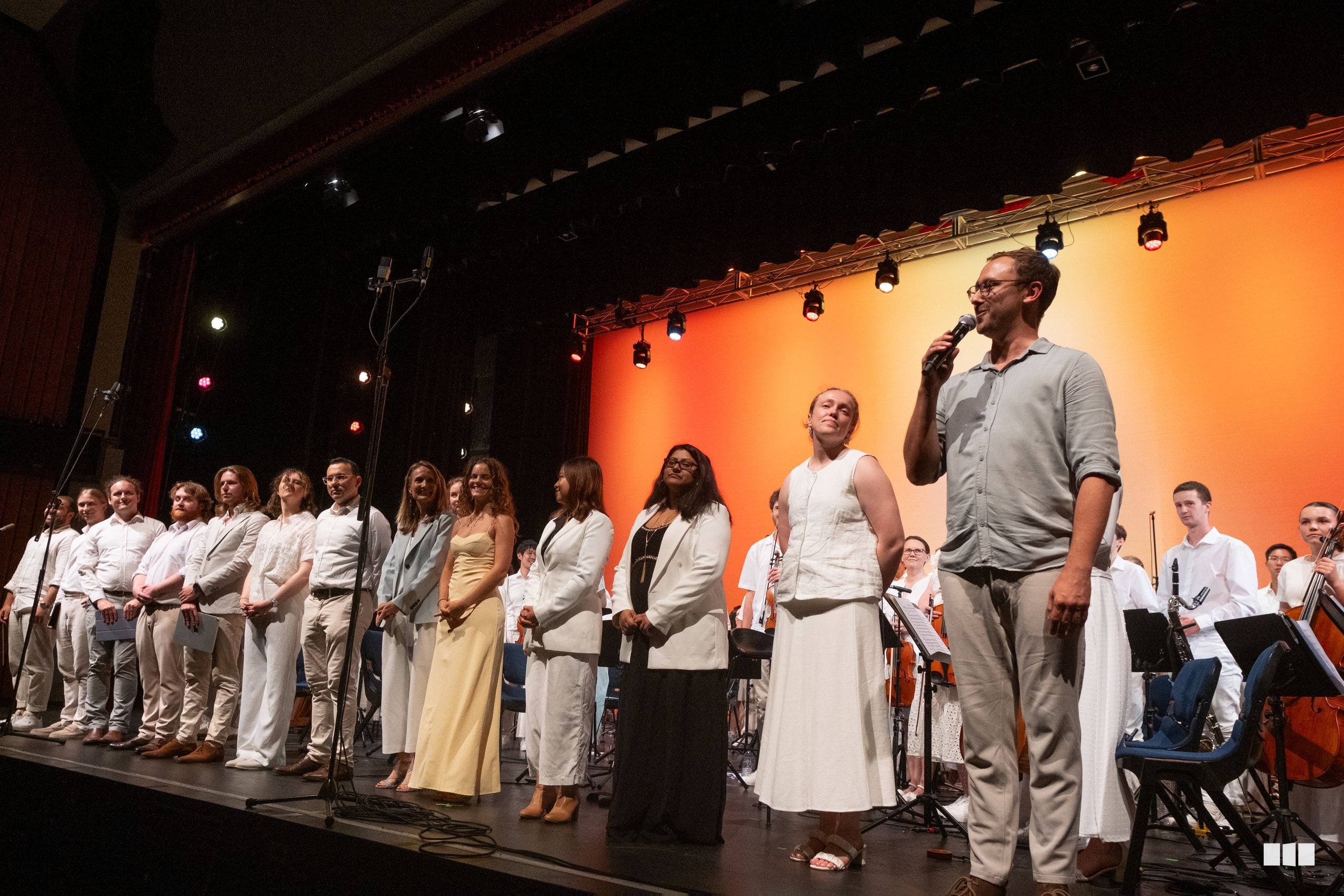 Orchestra and choir members stand together on stage during a performance with warm orange lighting and professional staging.