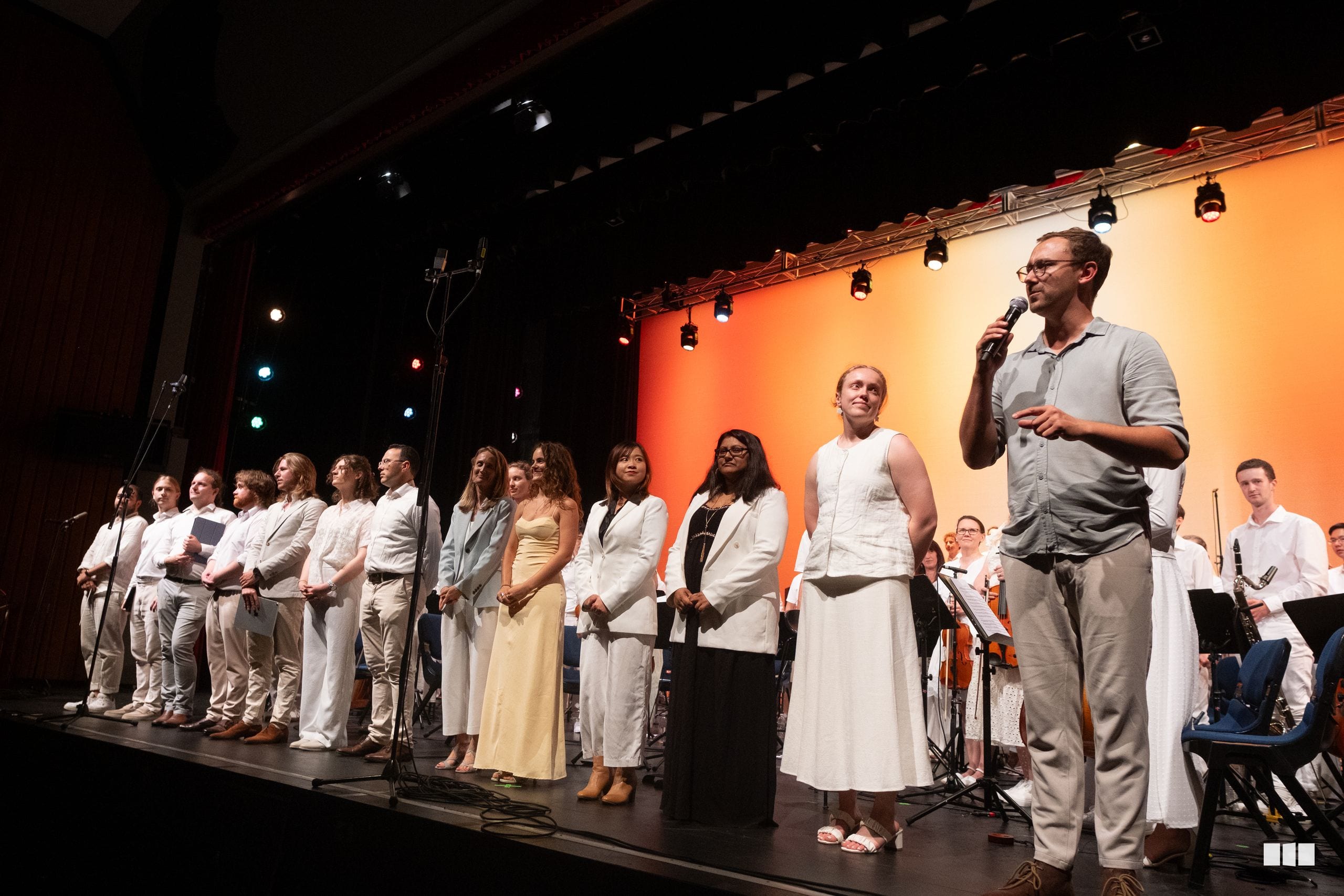 A conductor addresses the ensemble on stage while choir and orchestra members stand together during a performance or rehearsal.