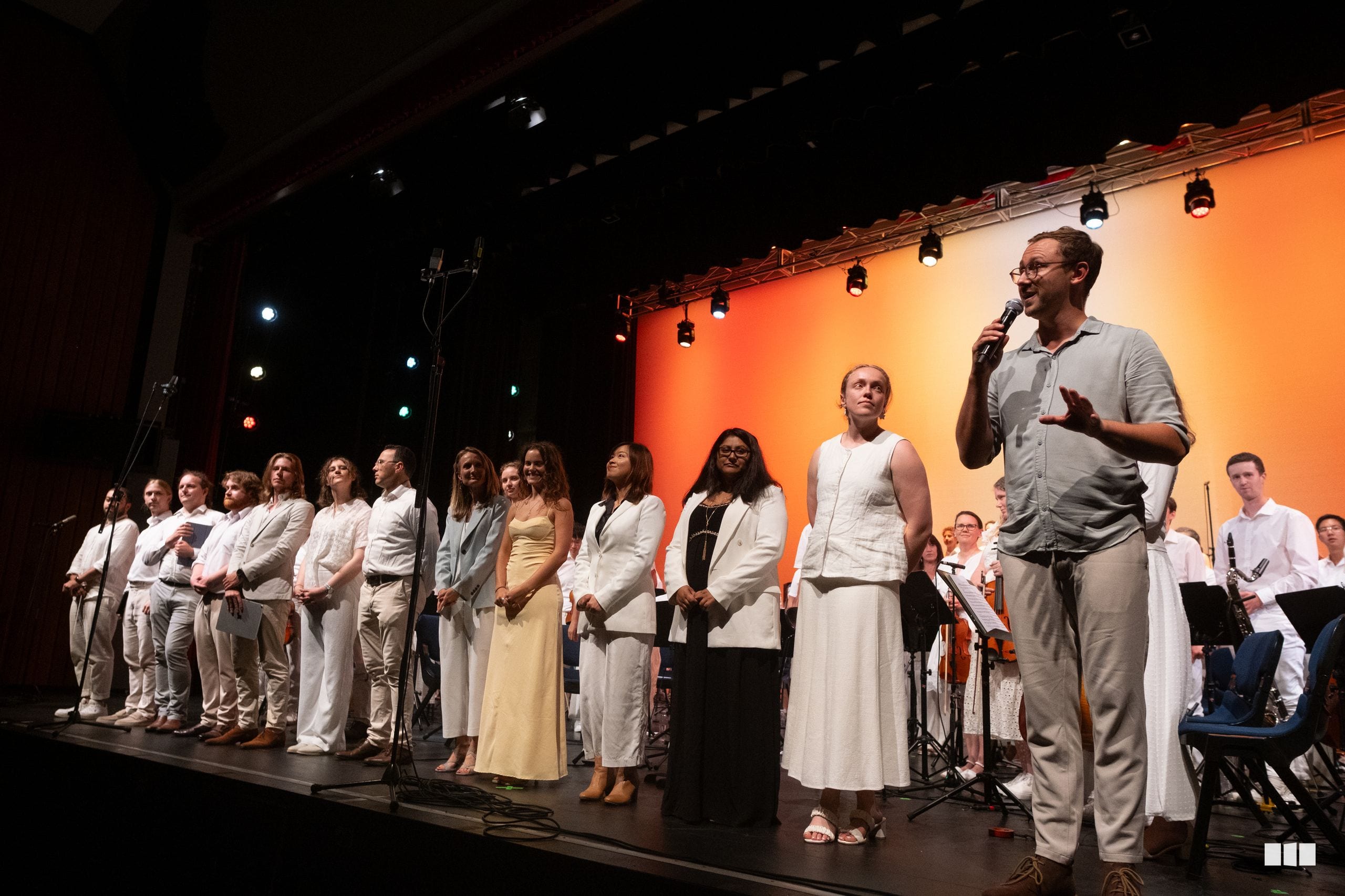 A conductor addresses the ensemble on stage while choir and orchestra members stand together during a performance.