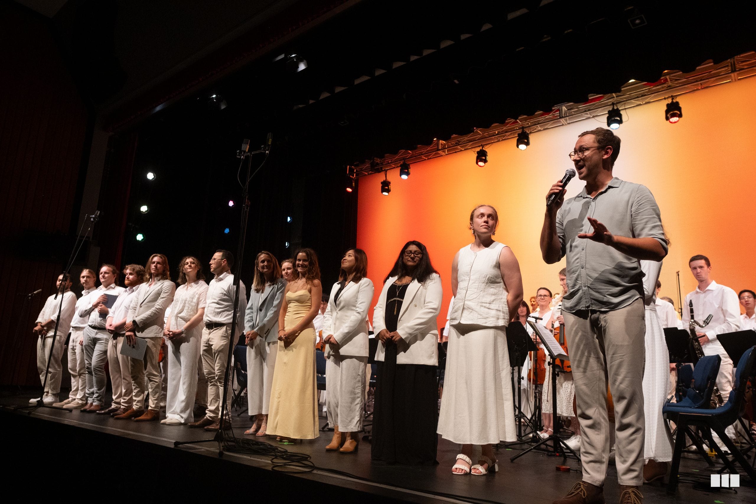A conductor addresses the choir and orchestra on stage during a performance with warm orange backlighting.