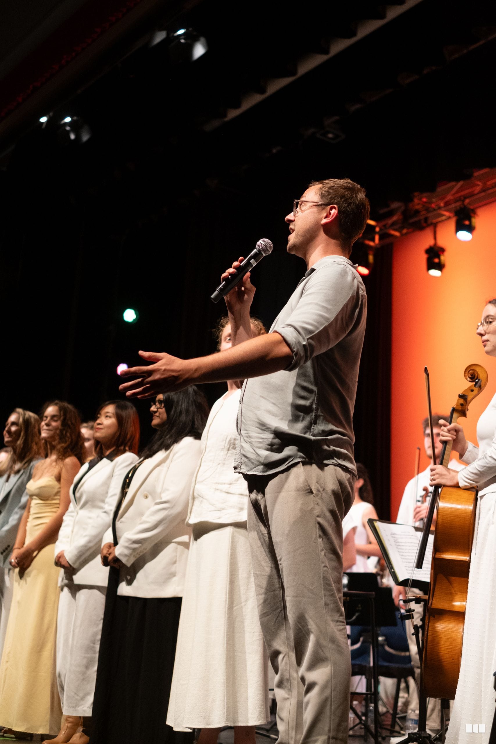 A male soloist sings into a microphone while addressing the audience, accompanied by orchestra musicians and choir members on stage.