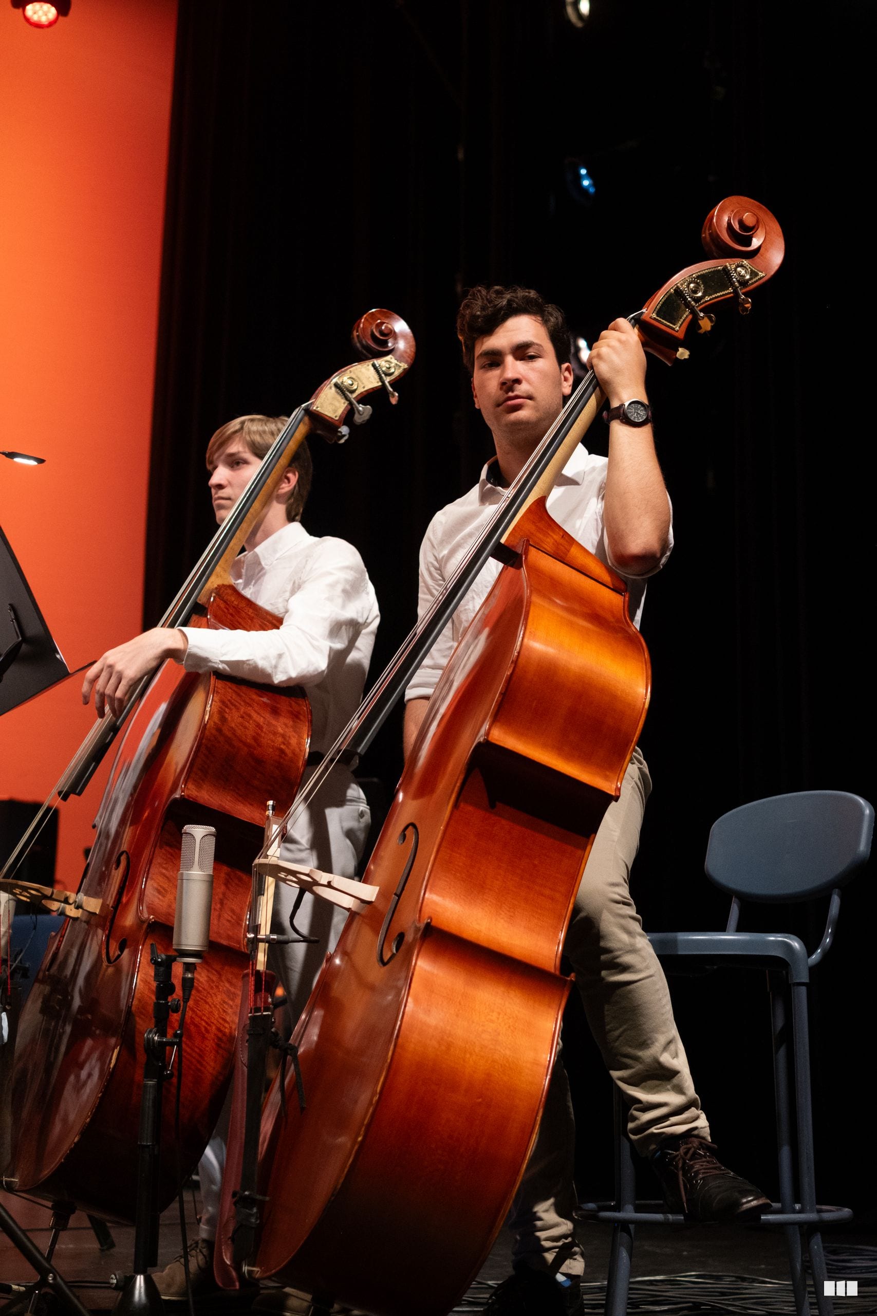 Two cellists perform together on stage, with the younger player on the left and an adult soloist on the right.