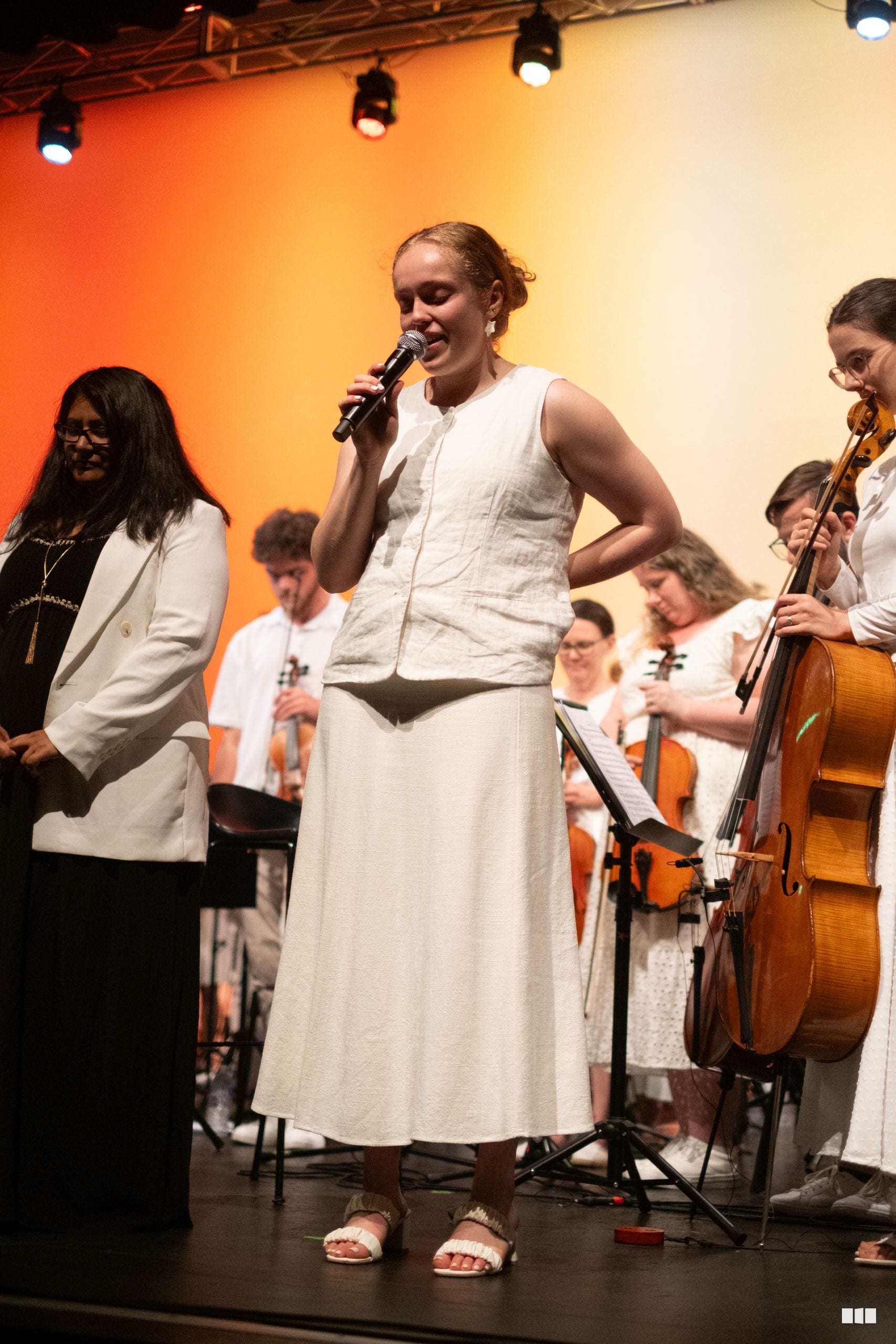 A soloist sings into a microphone while accompanied by orchestra members holding string instruments on stage under warm stage lighting.