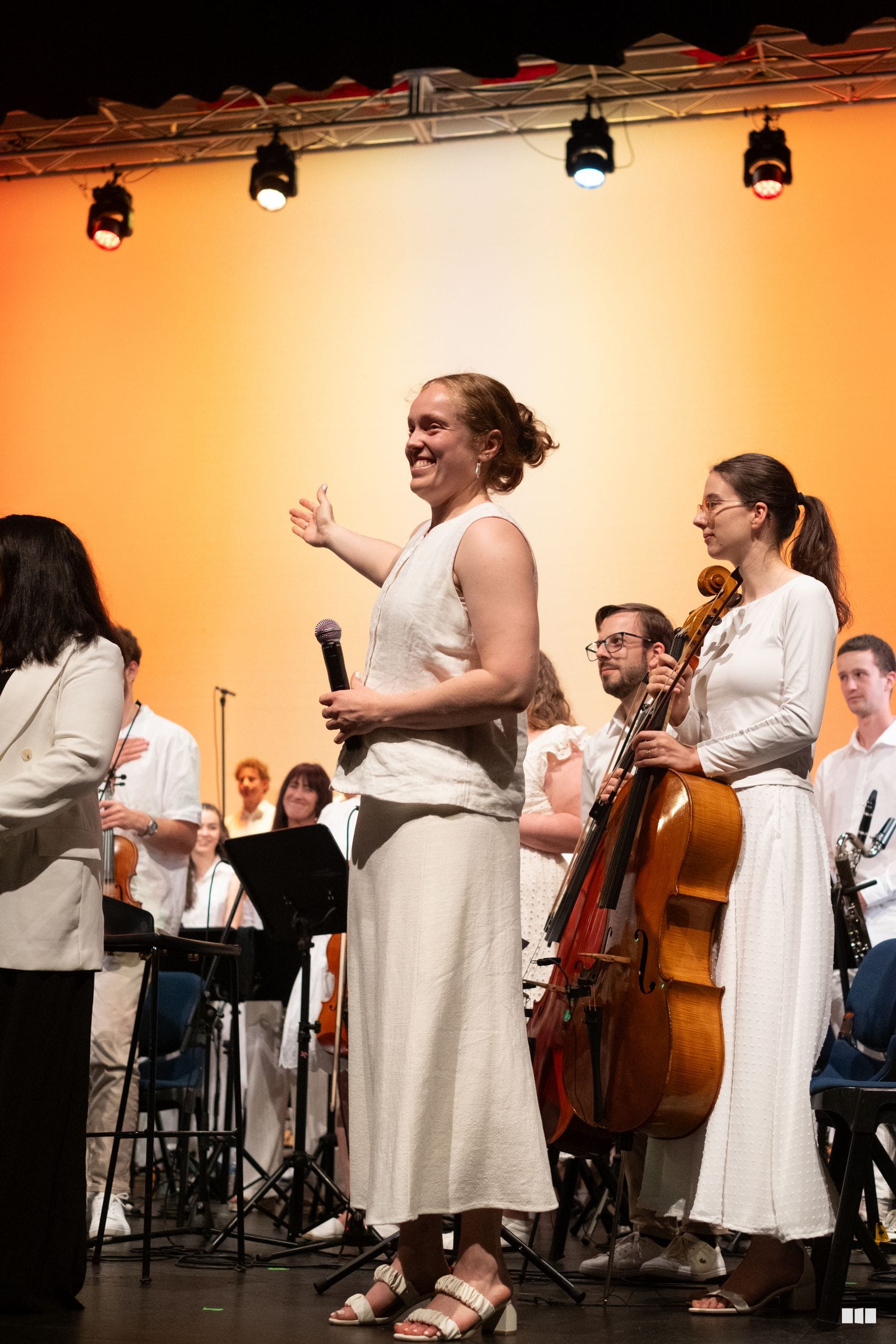 A female soloist speaks to the audience while holding a microphone, accompanied by orchestra members in white attire on stage.