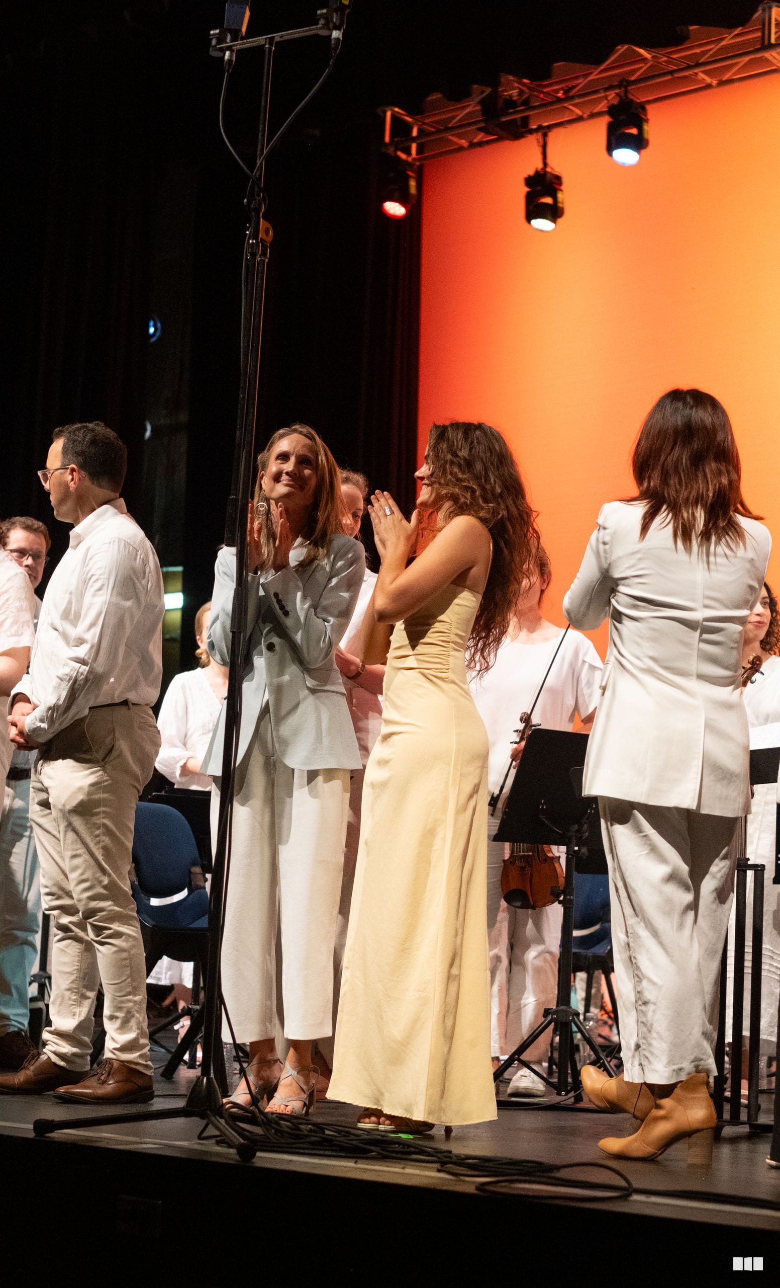 Choir members in light-colored formal attire stand together on stage during a performance, with warm orange stage lighting in the background.