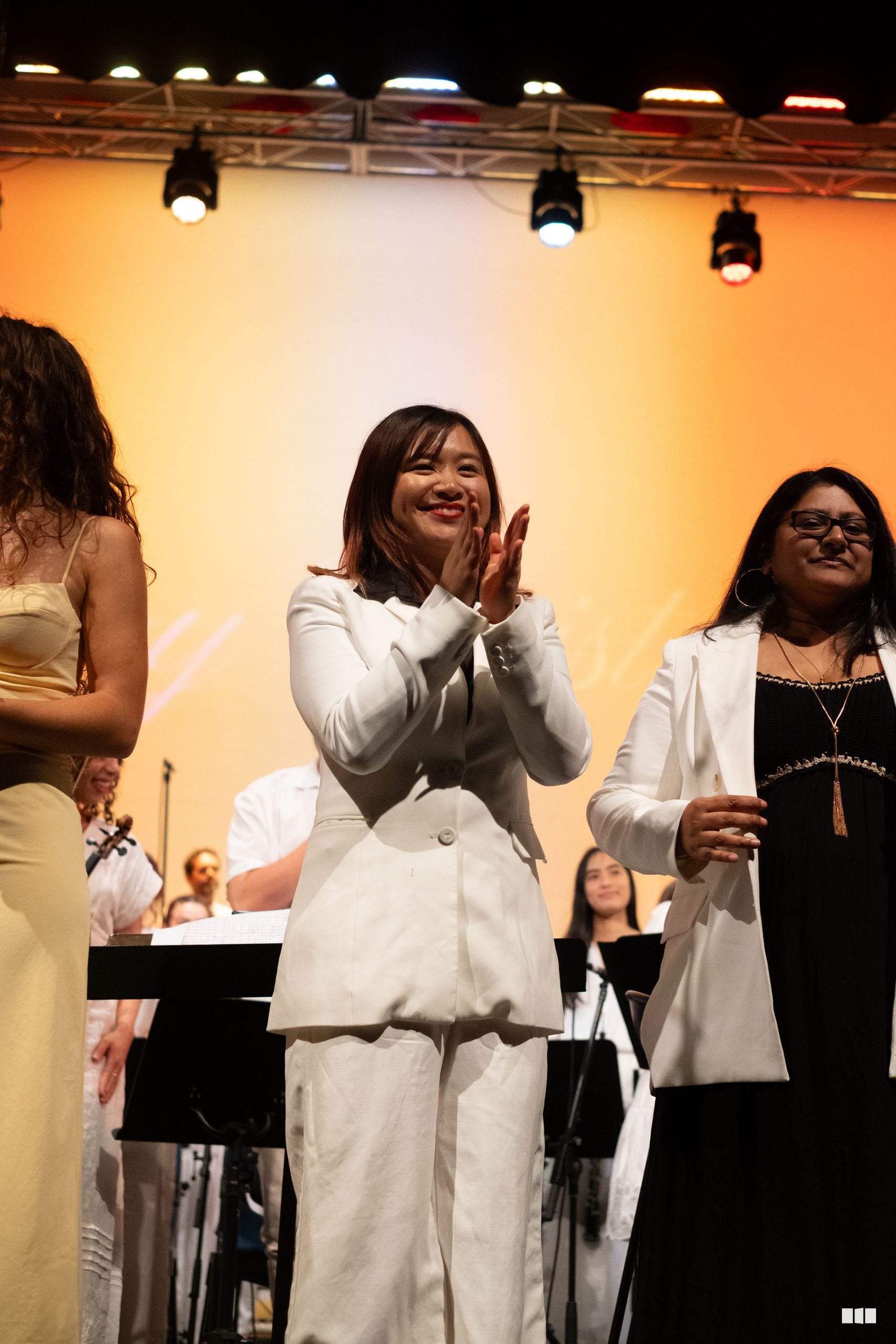 A woman in a white blazer and pants smiles and claps on stage while standing with colleagues in front of a warm-lit backdrop.