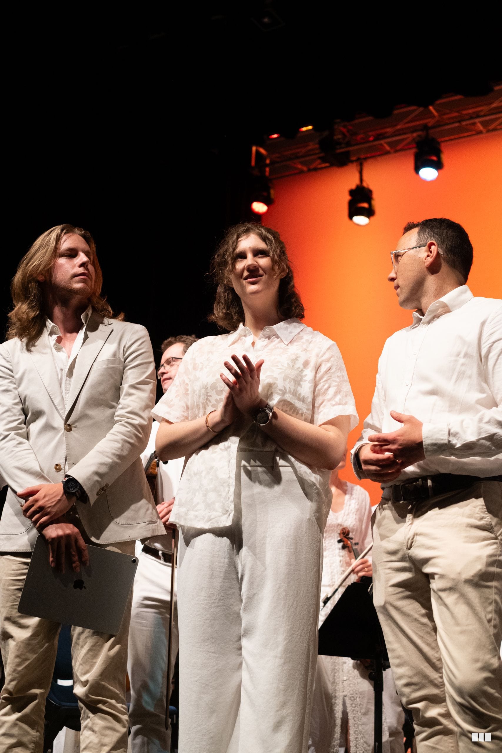 Three performers in light-colored clothing engaged in discussion on stage under warm orange lighting.