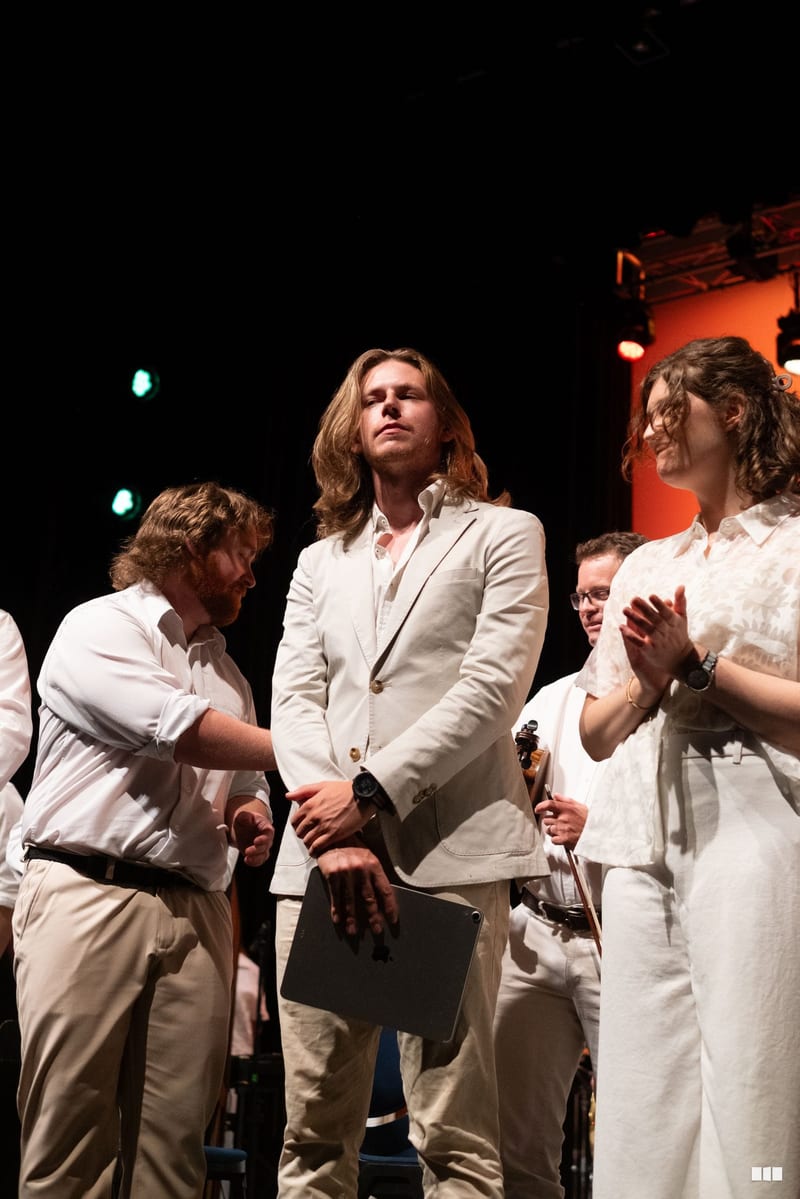 Several performers in cream and white formal attire stand together on stage during what appears to be a presentation or performance moment.