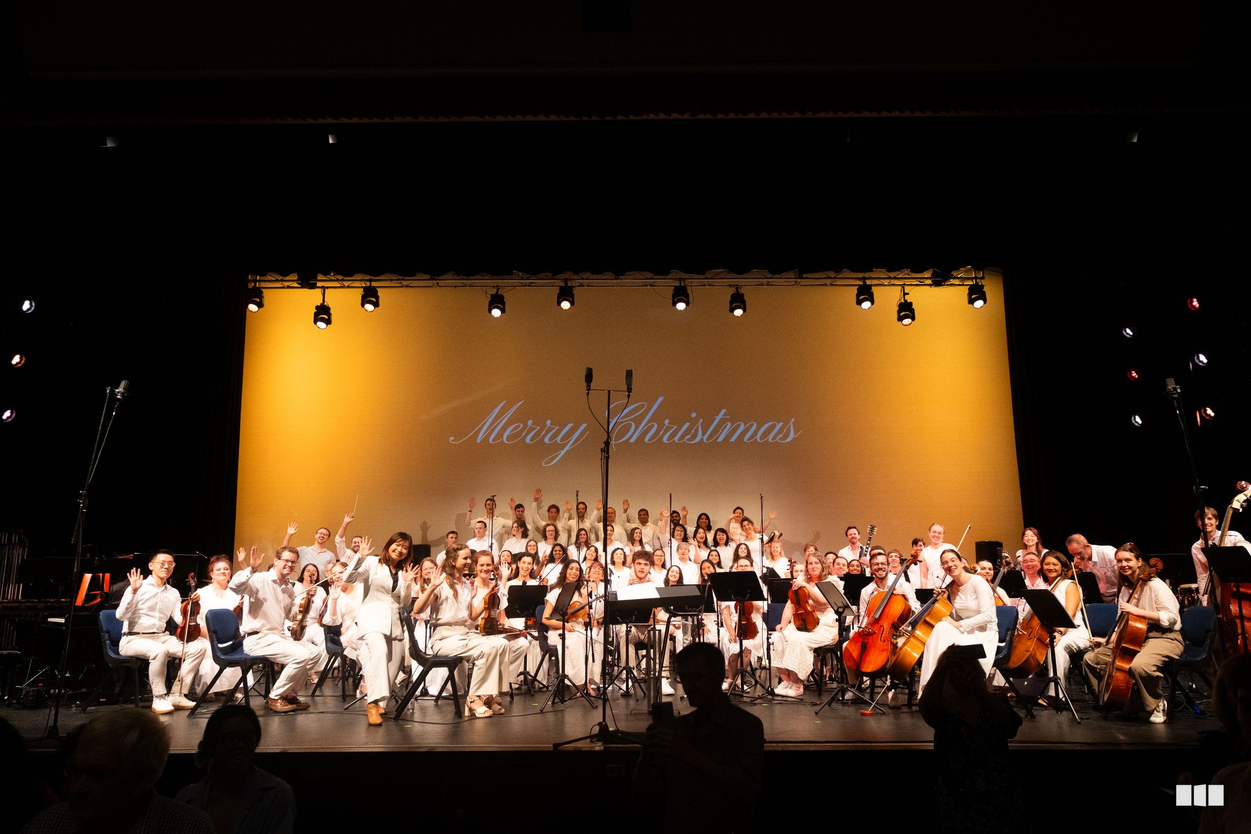 A large orchestra and choir perform a festive Christmas concert on stage under warm golden lighting with 'Merry Christmas' displayed on the backdrop.