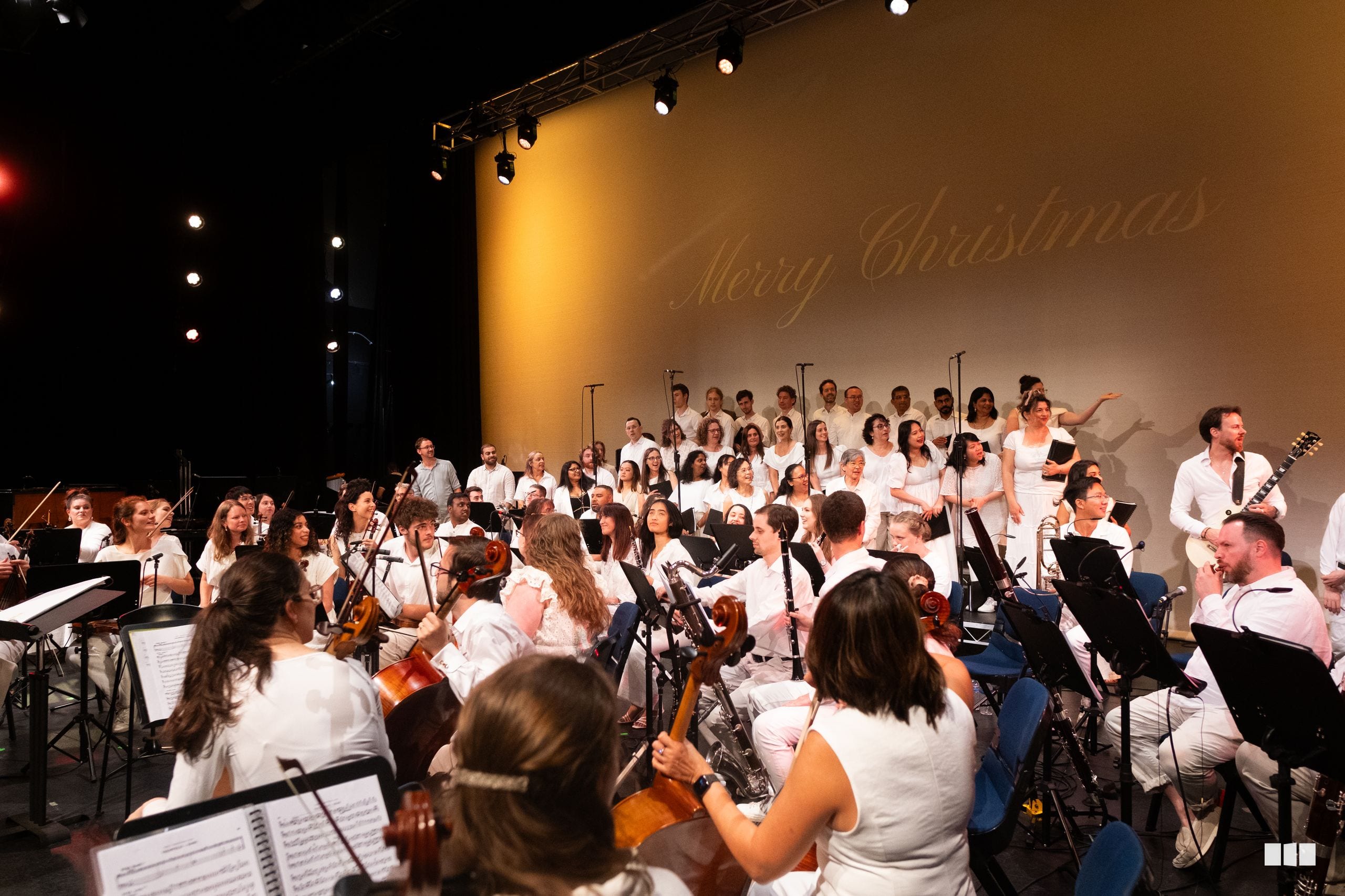 A large orchestra and choir in white attire perform together on stage with 'Merry Christmas' projected on the backdrop.
