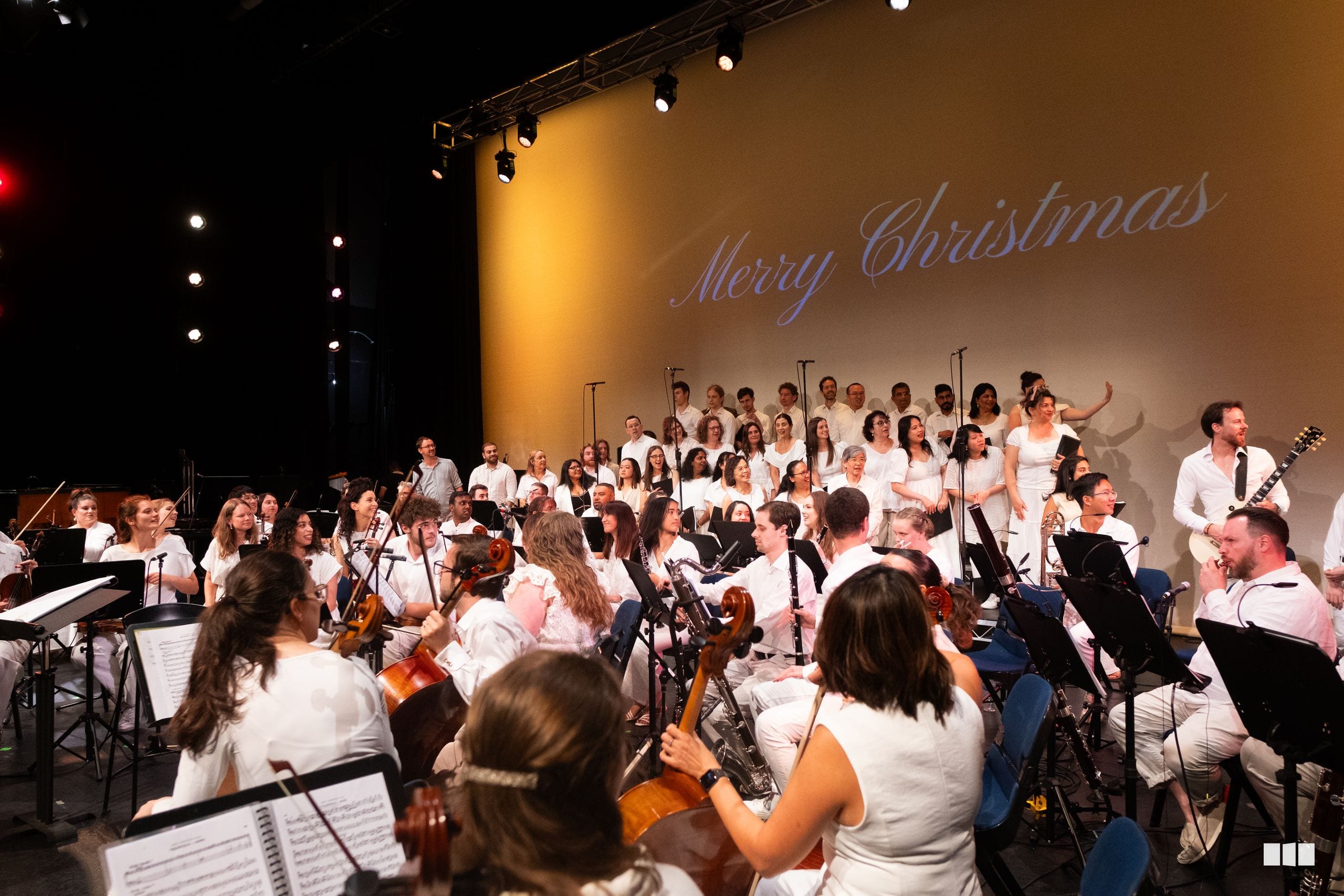 A large orchestra and choir perform a Christmas concert together on stage, with musicians in white and black attire and singers standing behind them under warm stage lighting.
