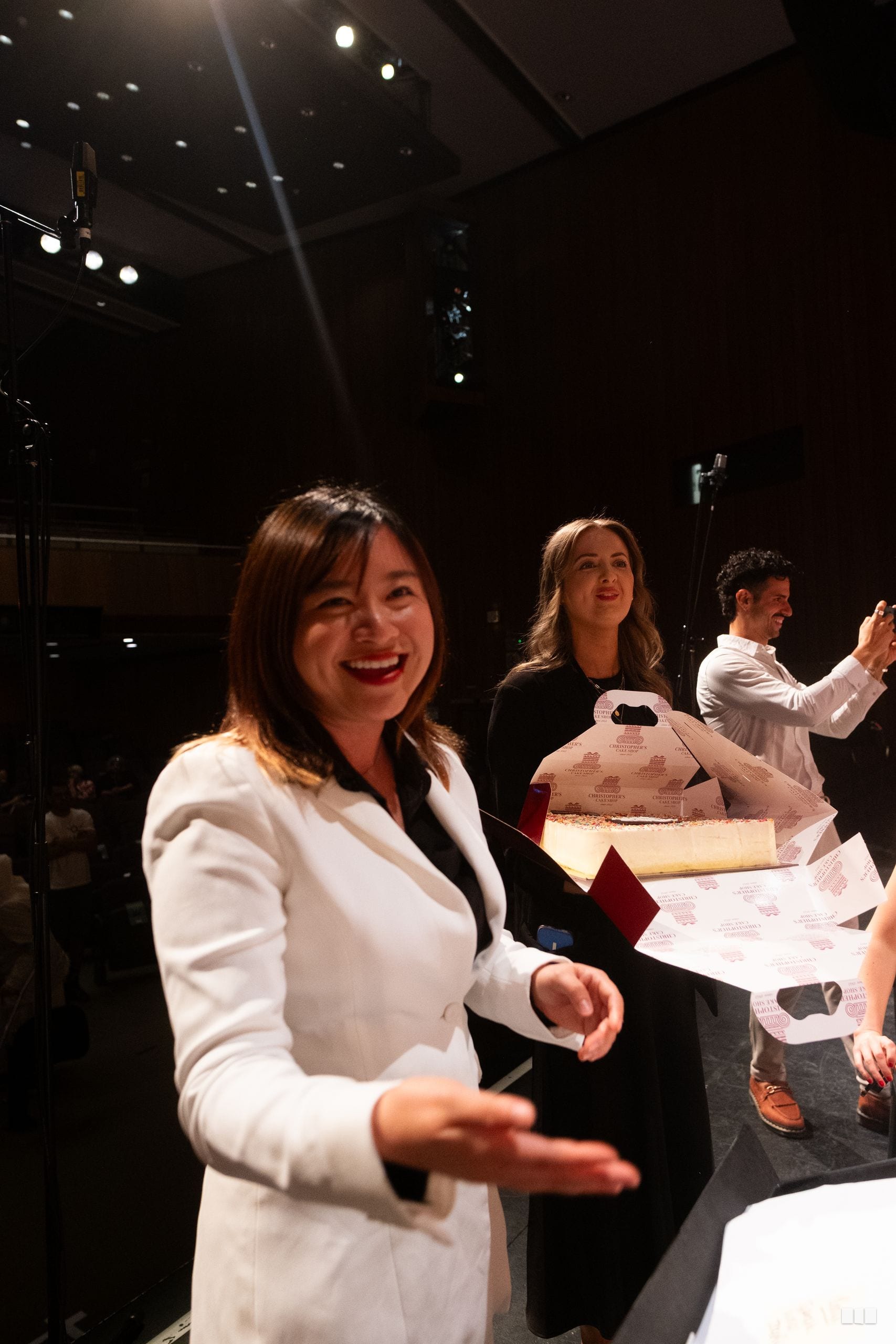 A woman in a white blazer smiles at the camera during what appears to be a formal event or rehearsal in a concert venue.