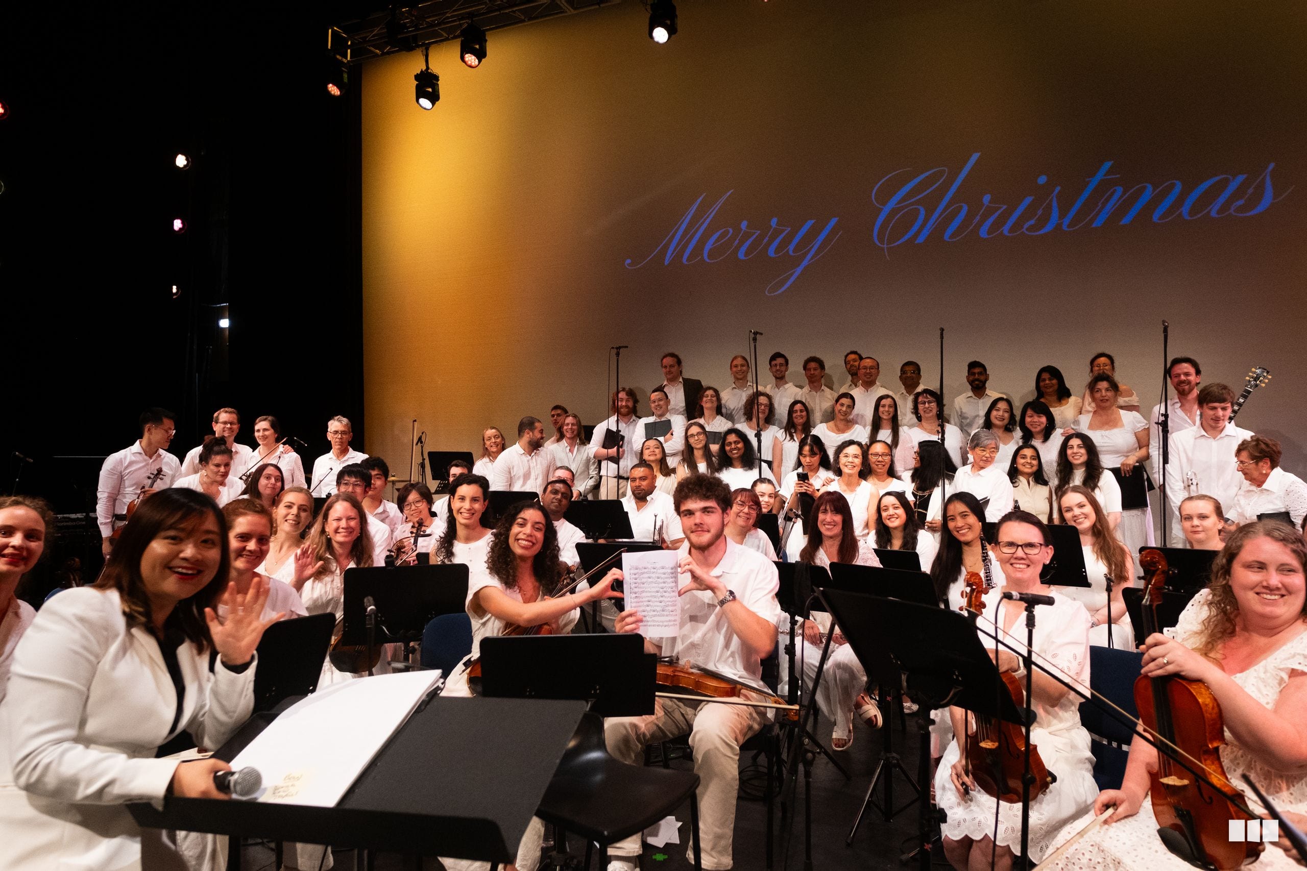 A large choir in white attire performs together on stage with 'Merry Christmas' displayed on the backdrop during a festive concert performance.