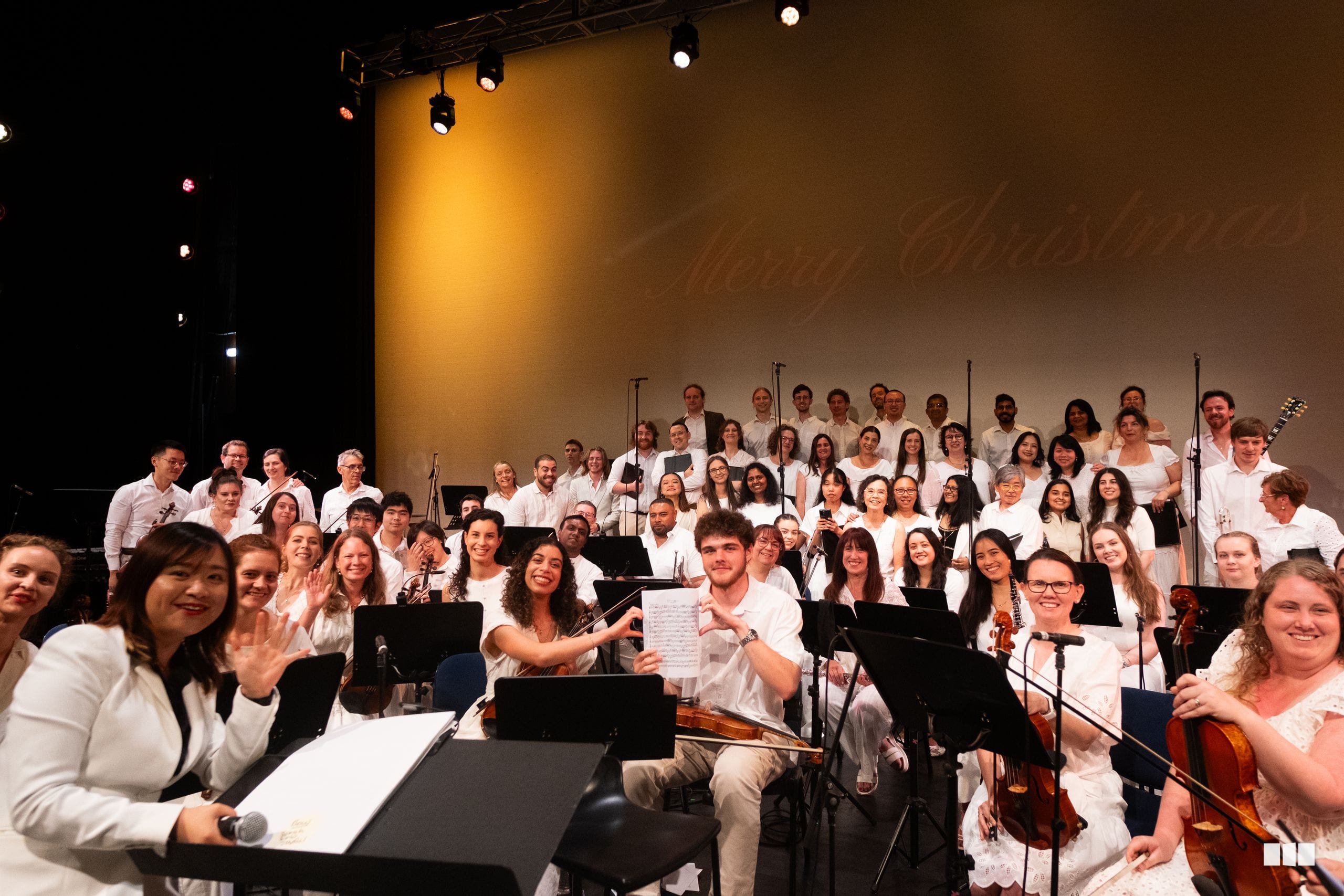 A large ensemble of orchestra and choir members dressed in white perform together on stage, smiling at the camera after their performance.