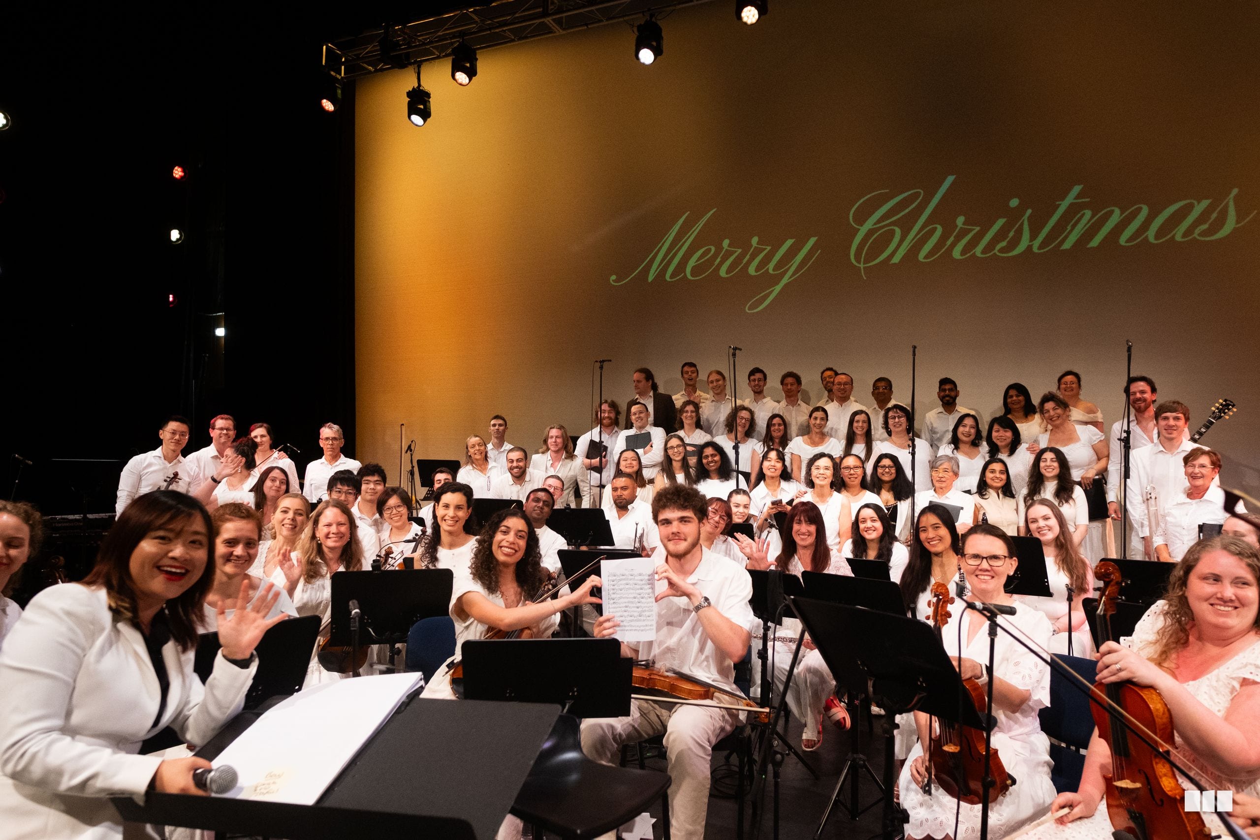 A large choir in white attire performs together on stage during a Christmas concert, with sheet music and music stands visible.