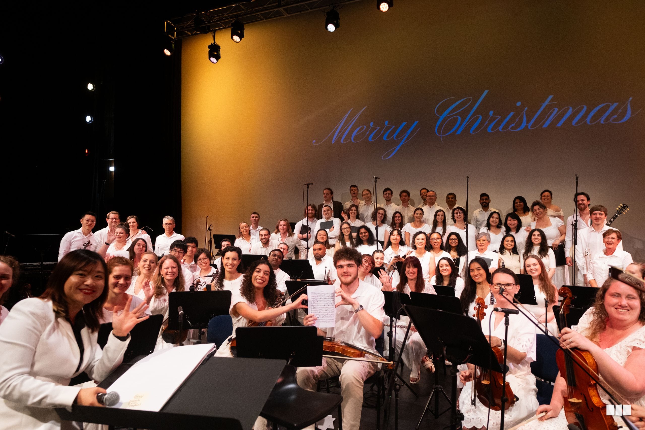 A large choir in white shirts performs on stage with a 'Merry Christmas' projection behind them, with orchestra members visible in the front rows.