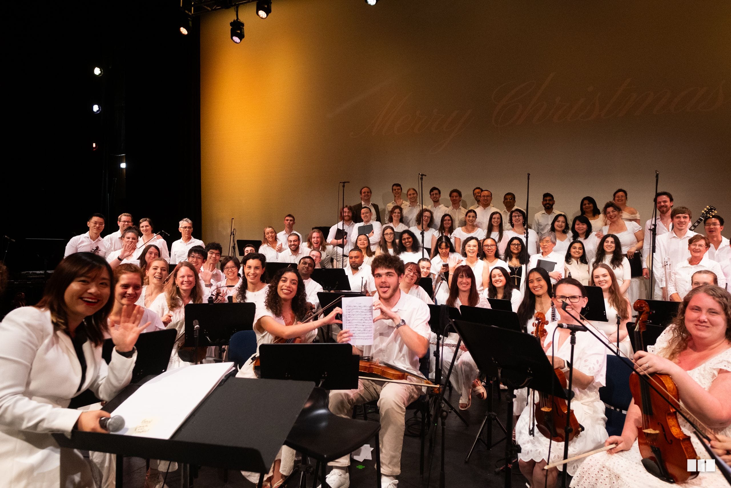 A large ensemble of orchestra and choir members in white attire perform together on stage during a concert.