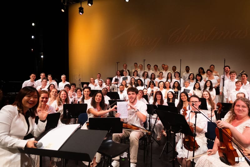 A large ensemble of orchestra and choir members in white attire perform together on stage during a concert.