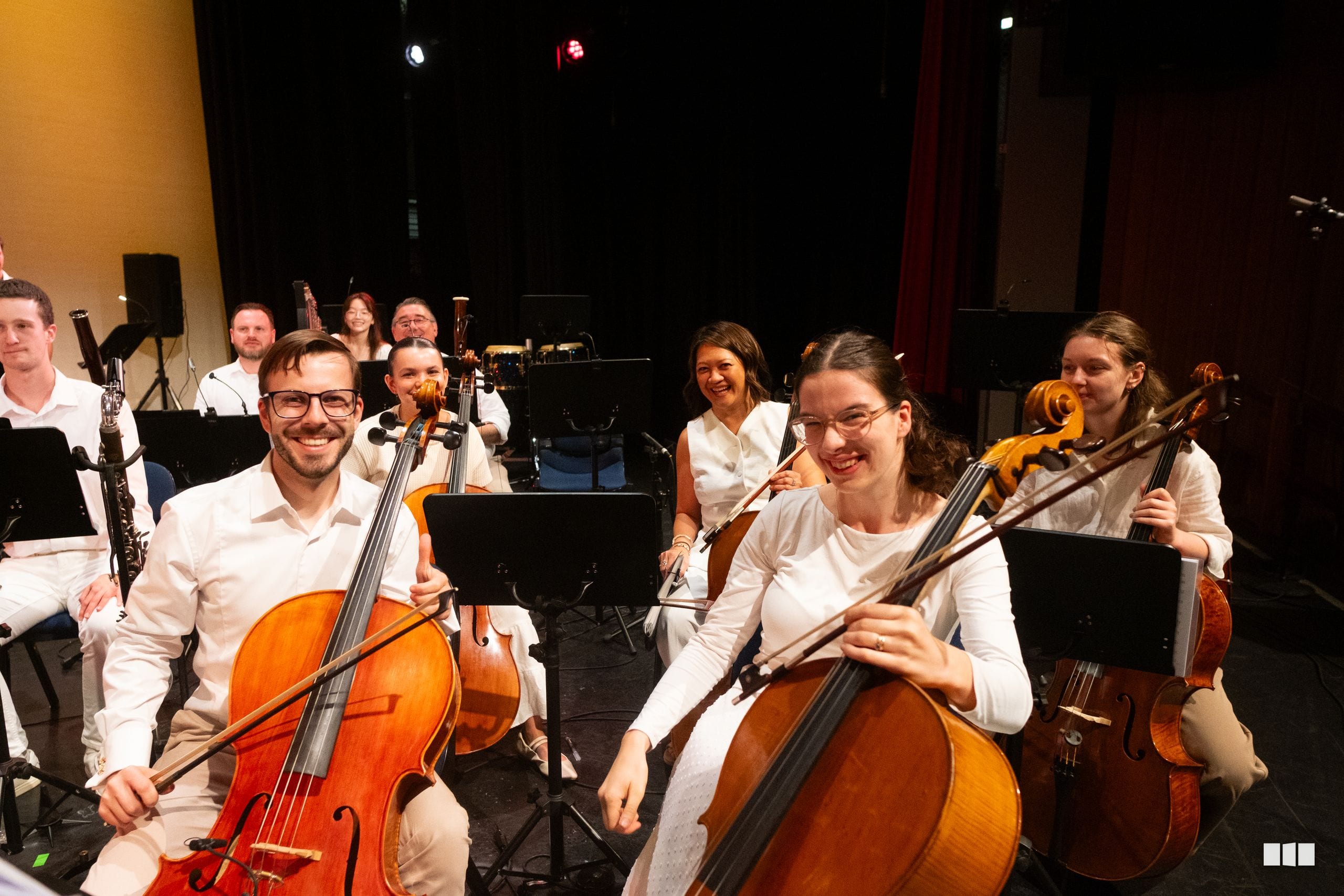 String section musicians in white attire perform together on stage with their instruments and sheet music.