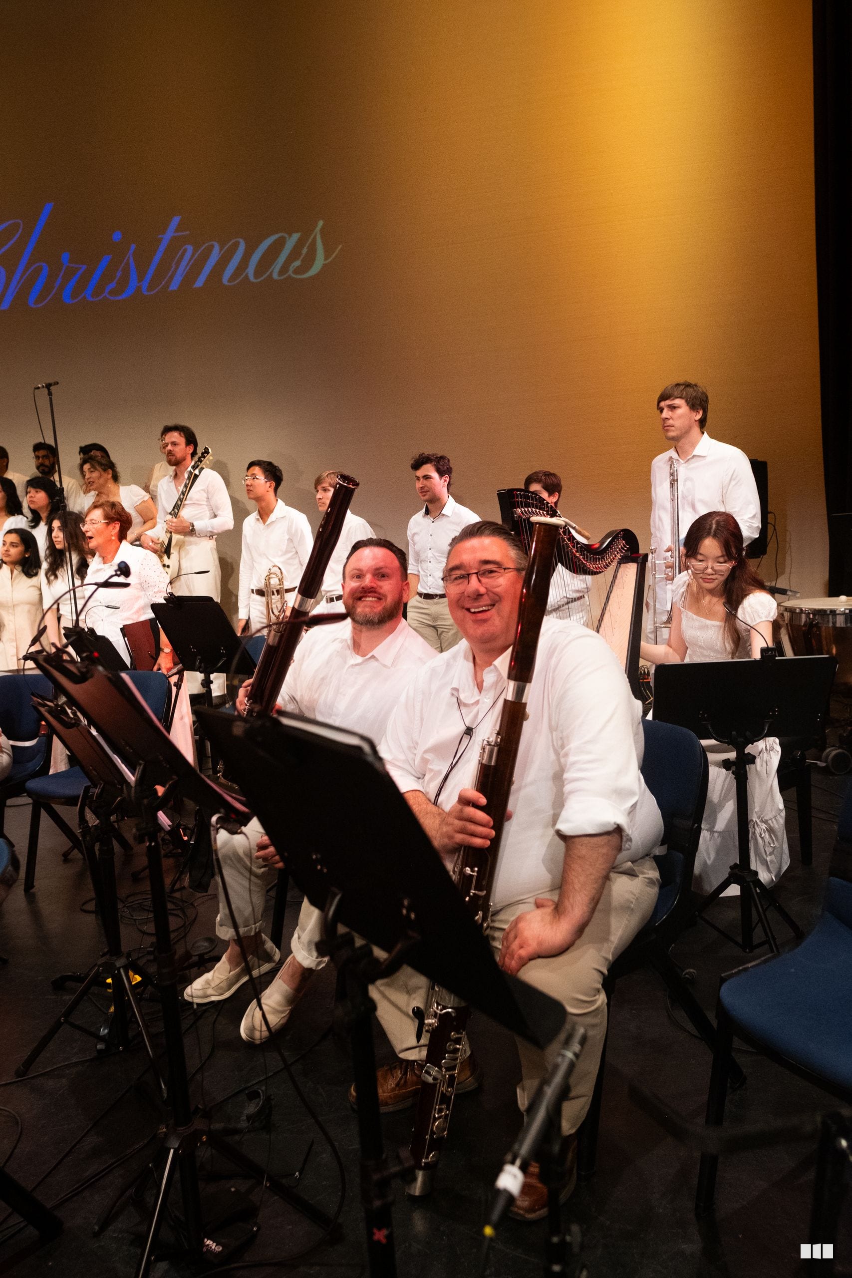 Two bassoon players smile at the camera during a Christmas concert performance with the full orchestra and choir assembled on stage behind them.