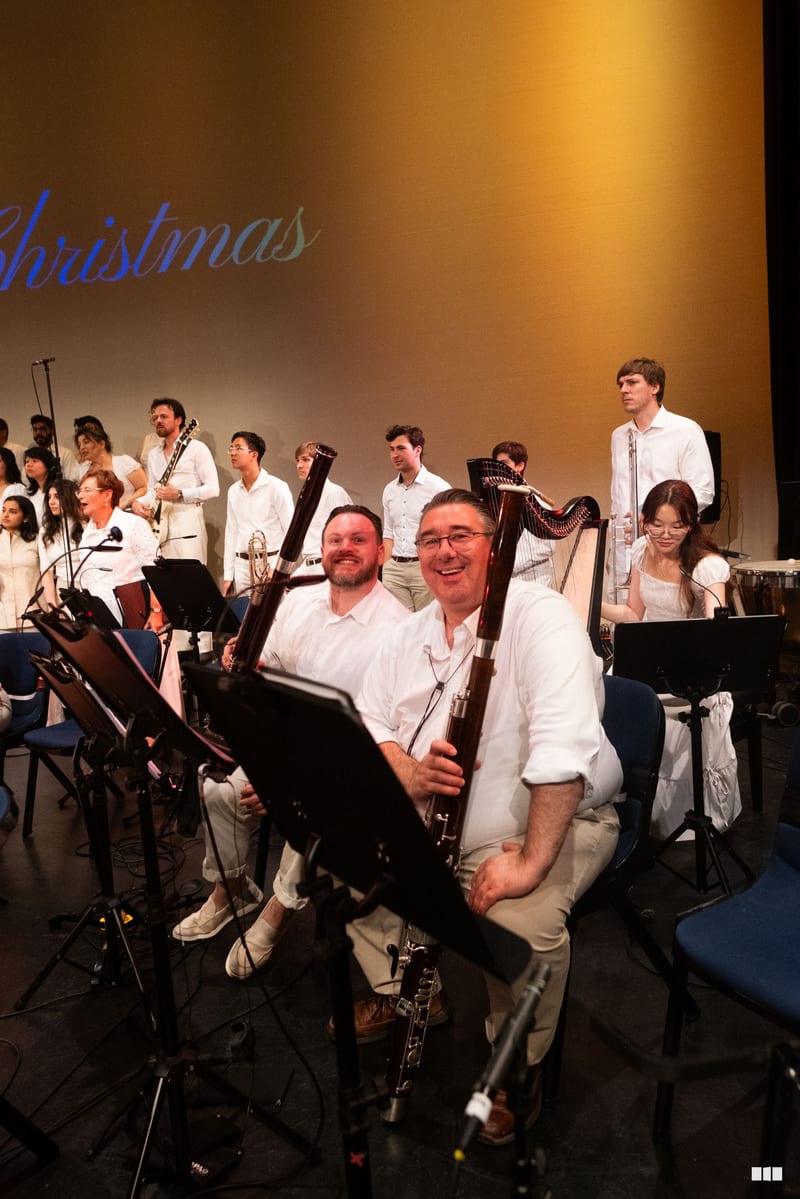 Two bassoon players smile at the camera during a Christmas concert performance with the full orchestra and choir assembled on stage behind them.