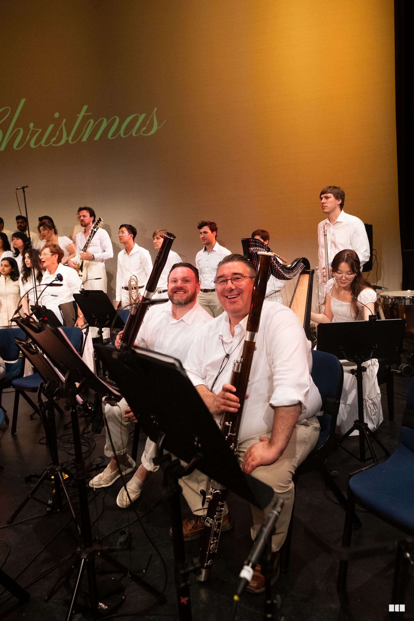 Orchestra members in white shirts smile together during a Christmas performance, with woodwind and string instruments visible on stage.