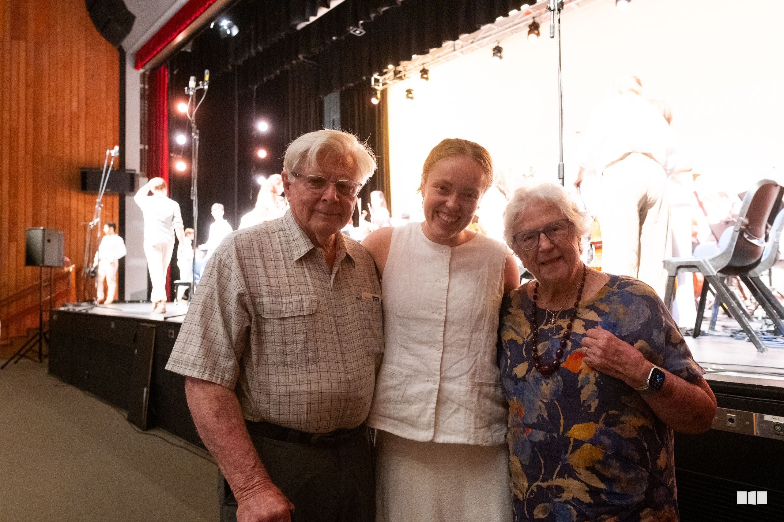 Three people pose together backstage at a concert venue with stage lighting and performers visible in the background.