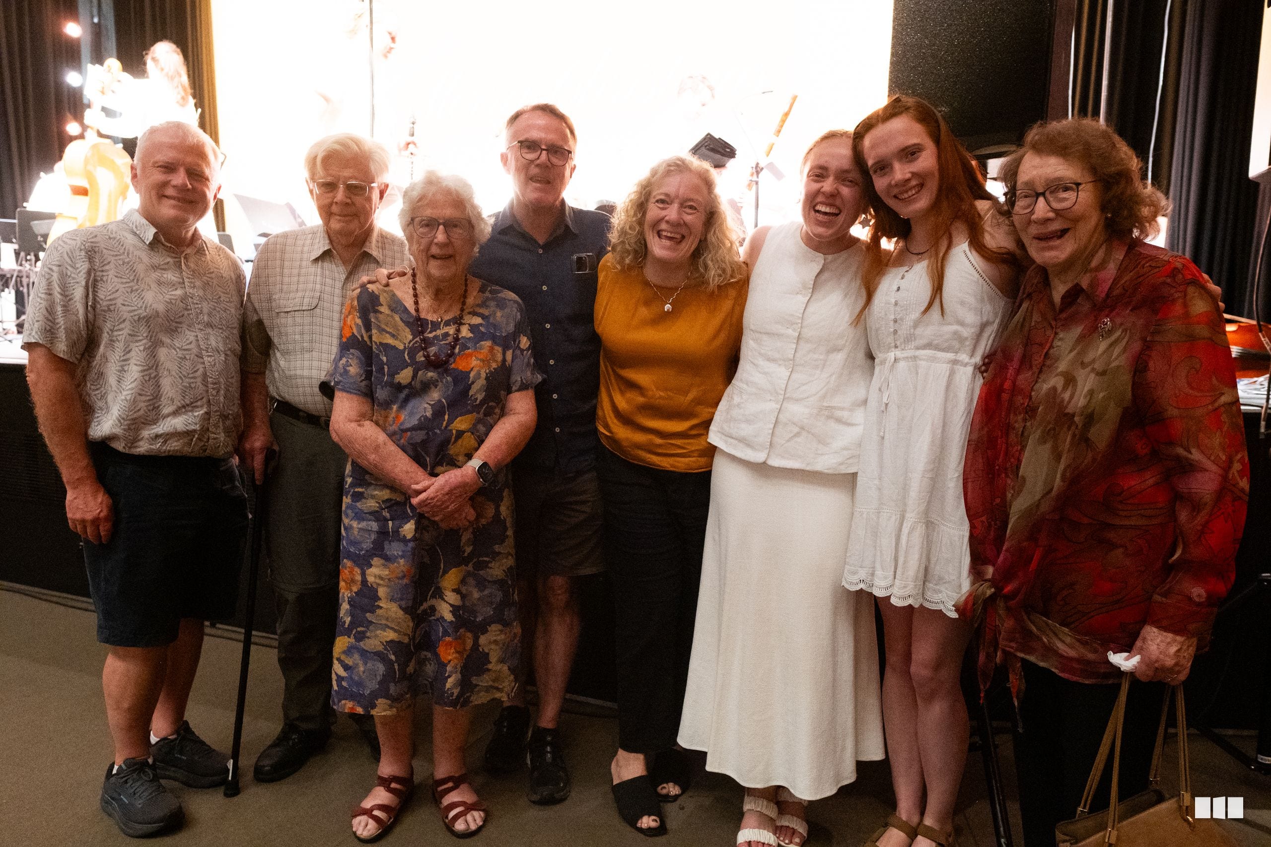 A group of nine people of mixed ages smile together at an indoor event venue, appearing to be at a social gathering or celebration.