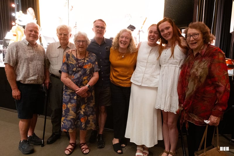 A group of nine people of mixed ages smile together at an indoor event venue, appearing to be at a social gathering or celebration.