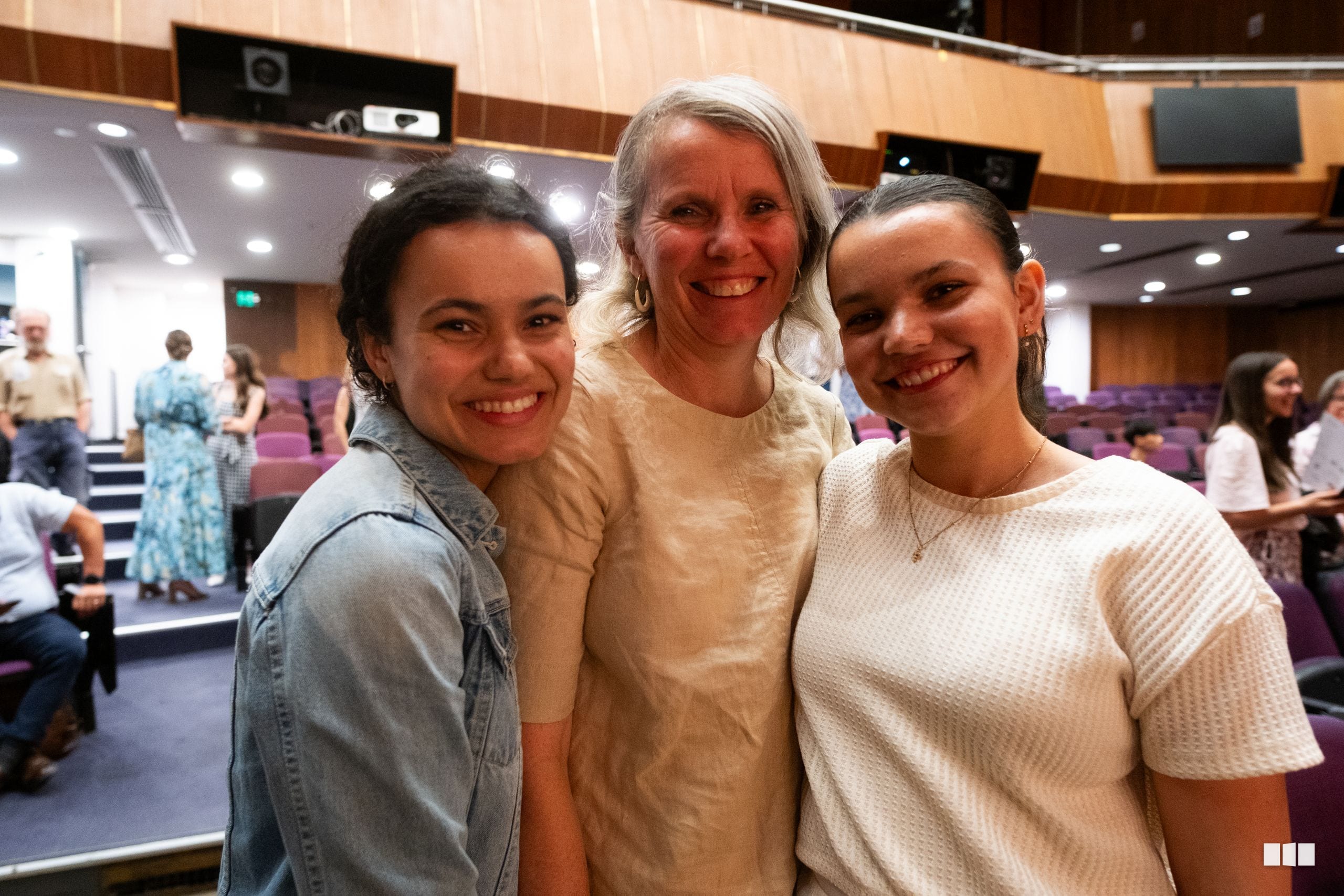 Three smiling women pose together in an auditorium during a community gathering event.