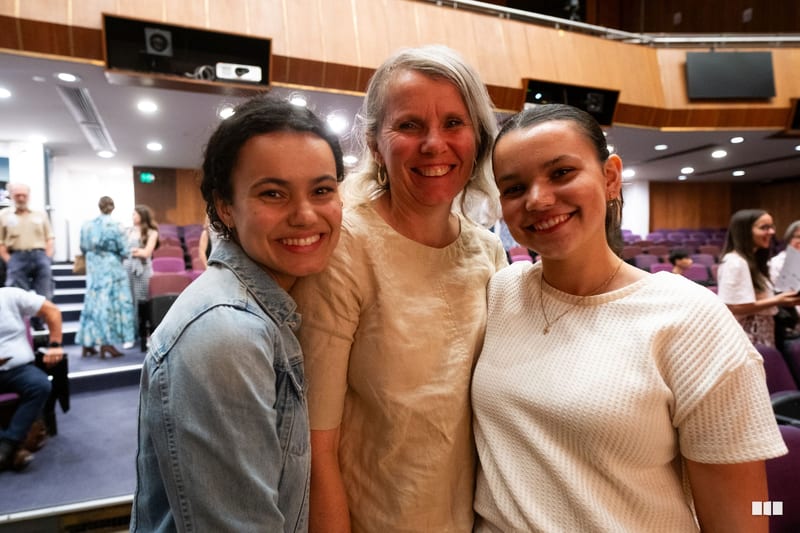 Three smiling women pose together in an auditorium during a community gathering event.
