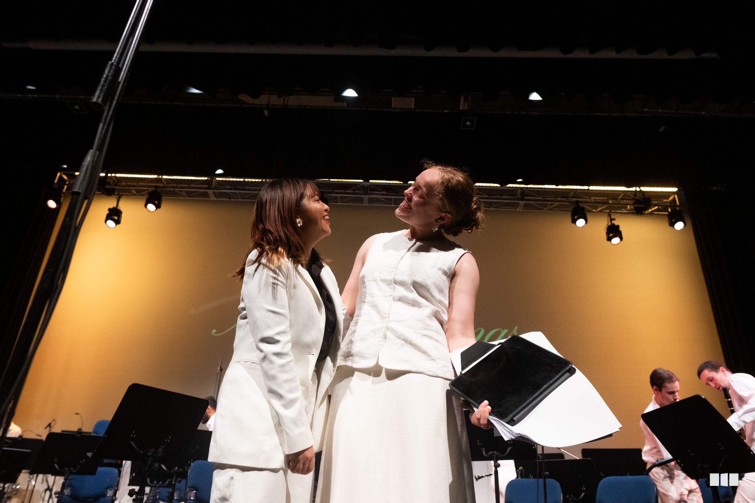 Two performers in white attire engage warmly on stage during a choral performance, with ensemble members visible in the background.