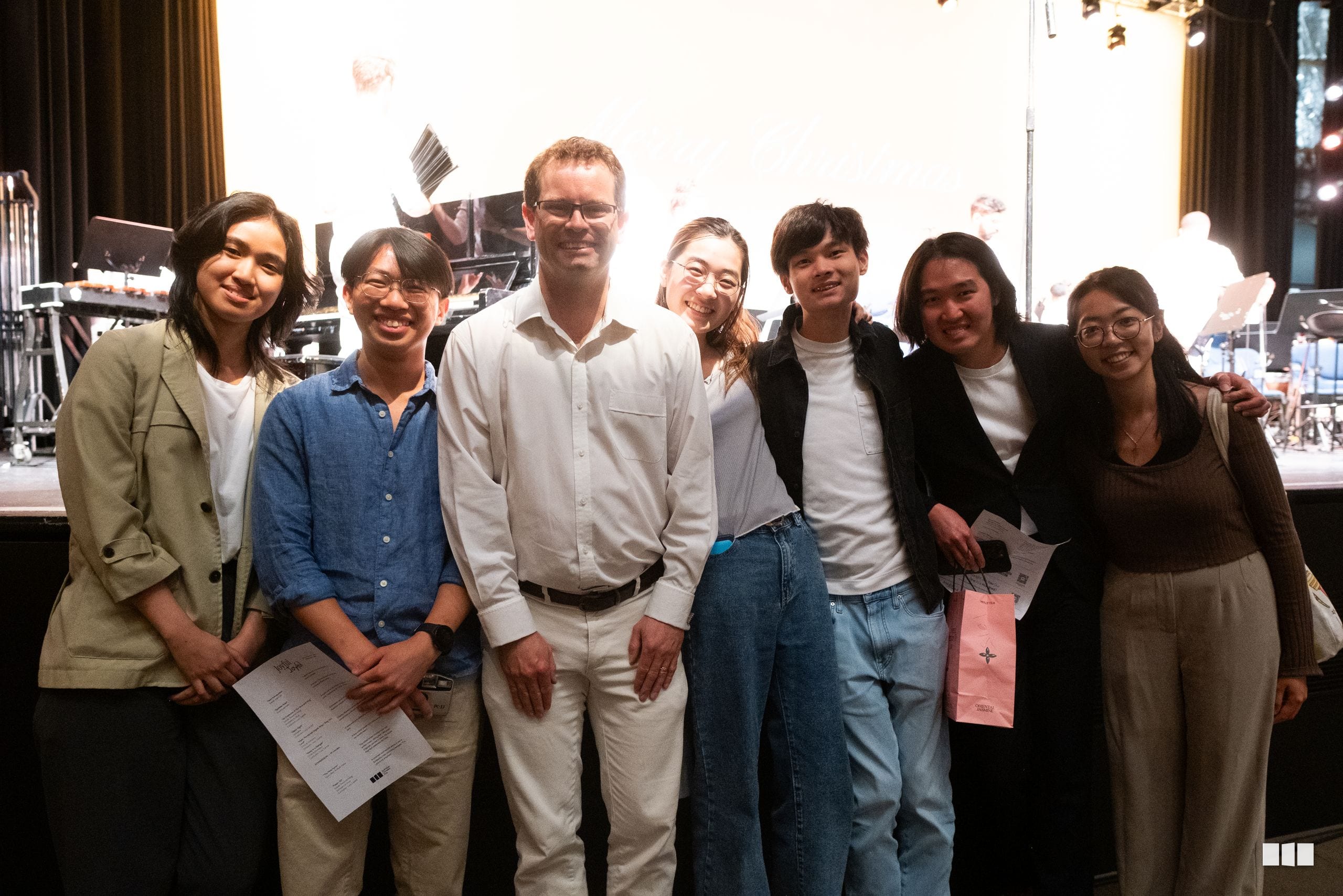 A diverse group of seven young people and one adult pose together on stage, smiling at the camera with musical equipment visible in the background.