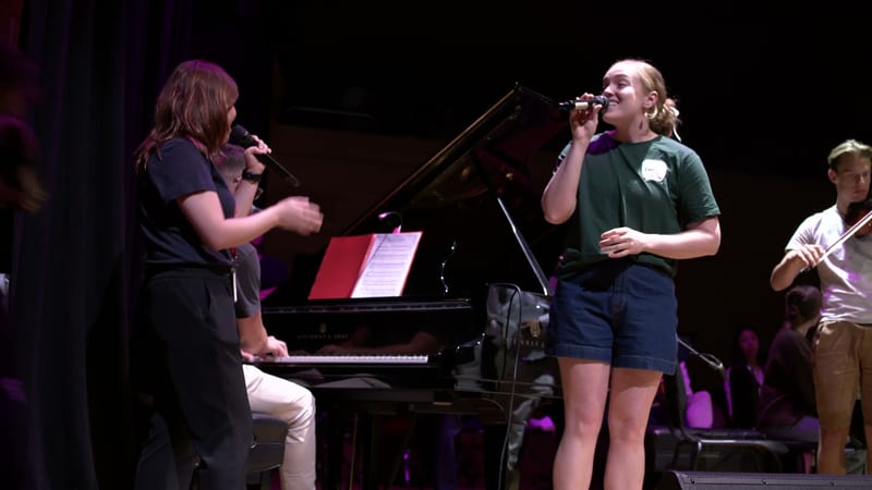 A young female soloist sings at the microphone while a pianist accompanies her on stage during a performance.