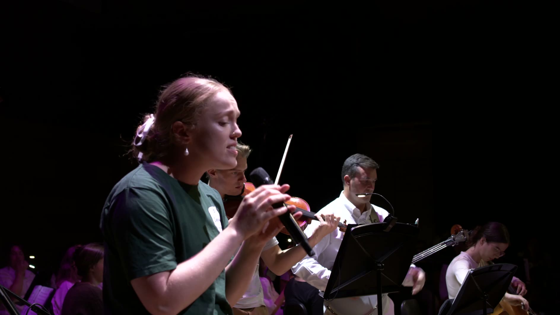 A young violinist performs with sheet music during an orchestral concert against a dark stage background.
