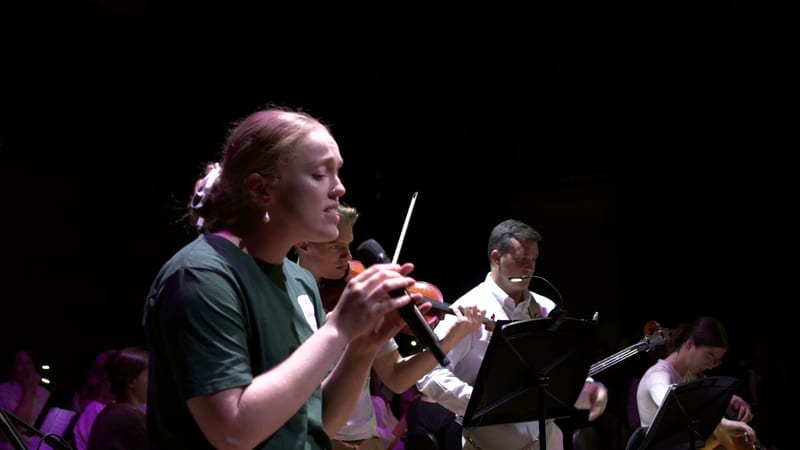 A young violinist performs with sheet music during an orchestral concert against a dark stage background.