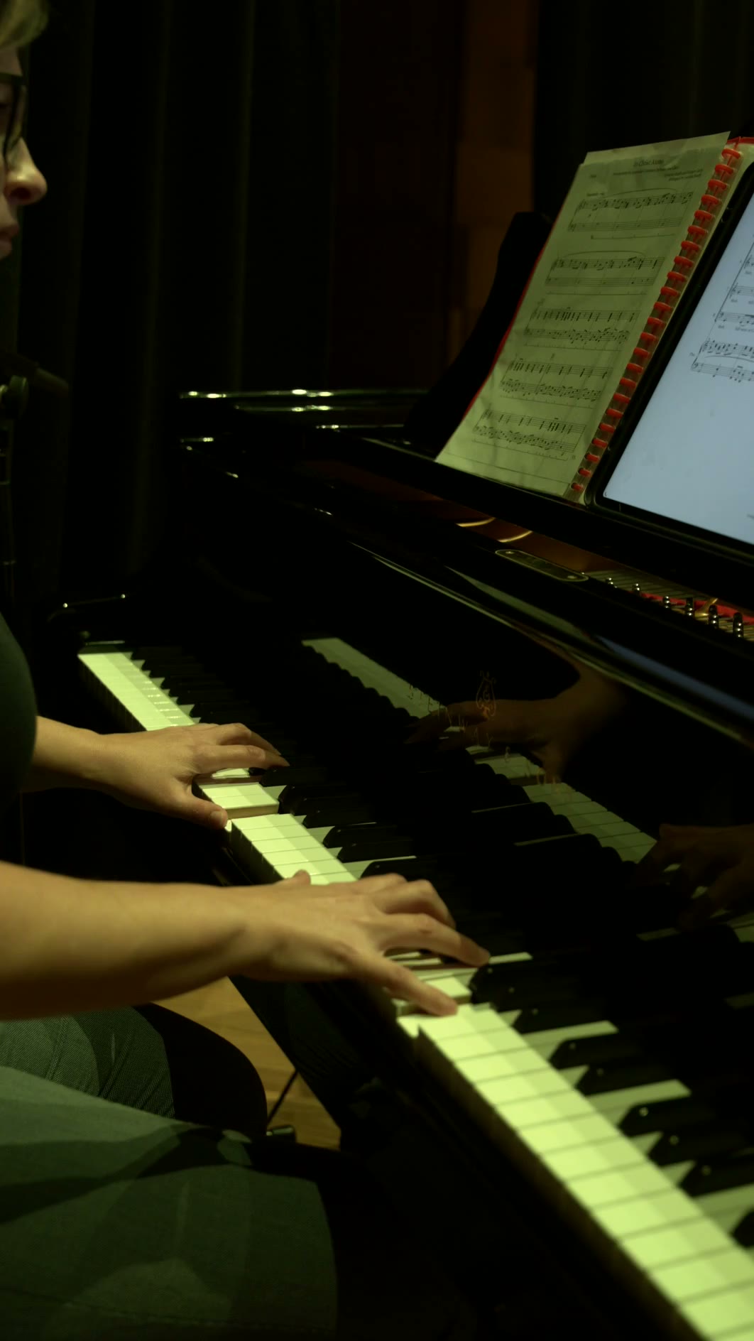 A young pianist's hands illuminate the keys during an evening performance, with sheet music visible on the music stand above.
