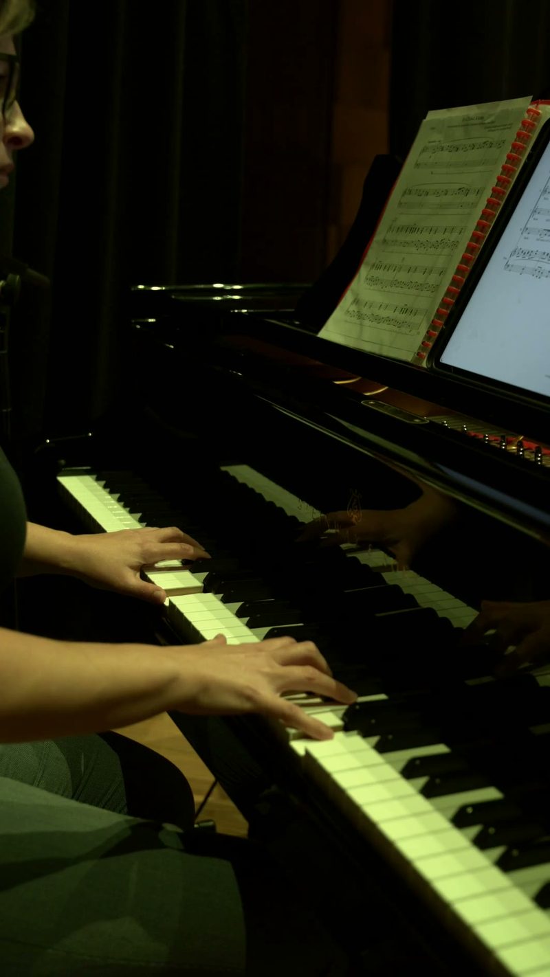 A young pianist's hands illuminate the keys during an evening performance, with sheet music visible on the music stand above.