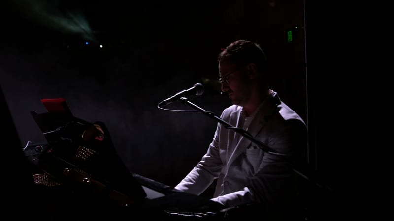 A musician performs at a keyboard while singing into a microphone during an evening concert performance.