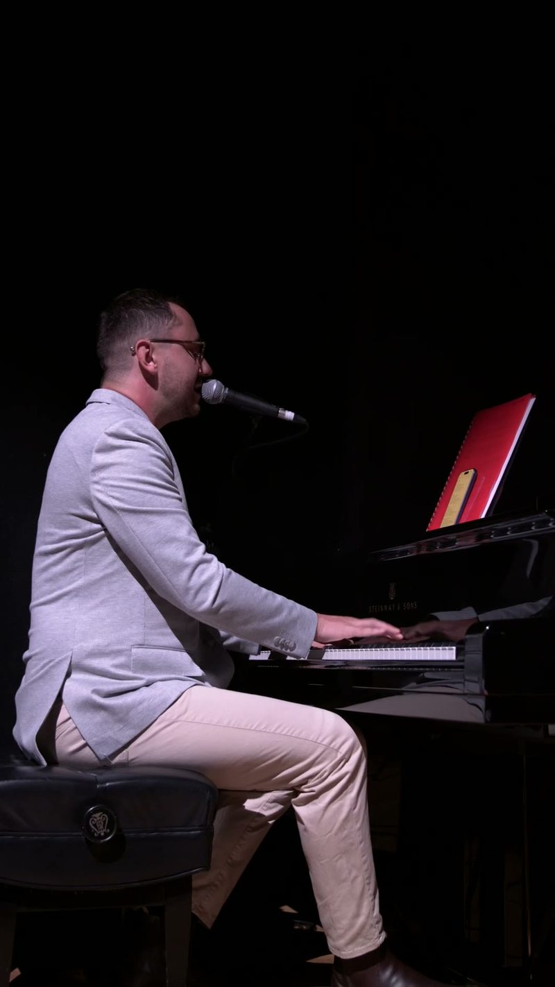 A musician in light-colored attire performs at the piano while singing into a microphone against a black background.