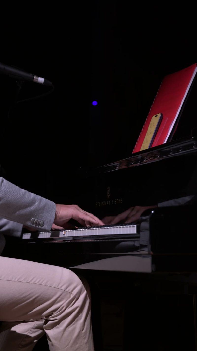 A pianist in formal attire performs at a grand piano illuminated against a dark background during an evening performance.