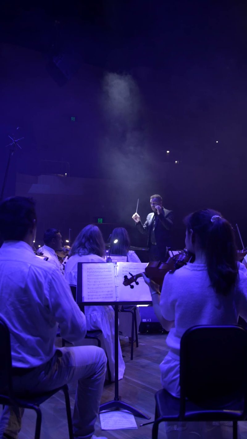 A conductor leads an orchestra during a performance with dramatic blue stage lighting and atmospheric effects.