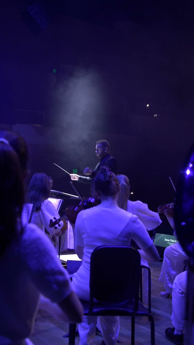 A conductor leads an orchestra during a performance with dramatic blue lighting and visible steam or fog effects on stage.