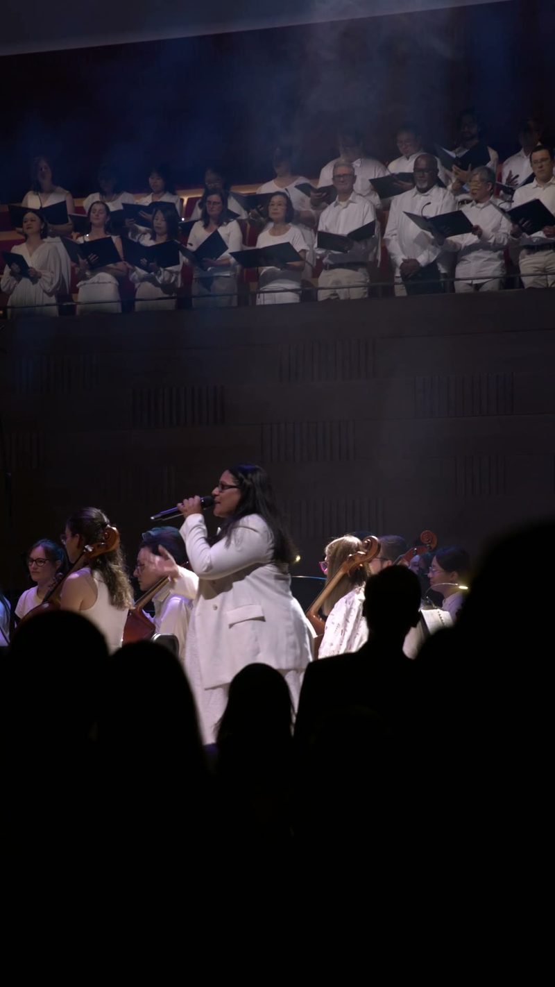 A soloist in white performs with microphone while a choir in white robes sings from the balcony above, accompanied by orchestra members.