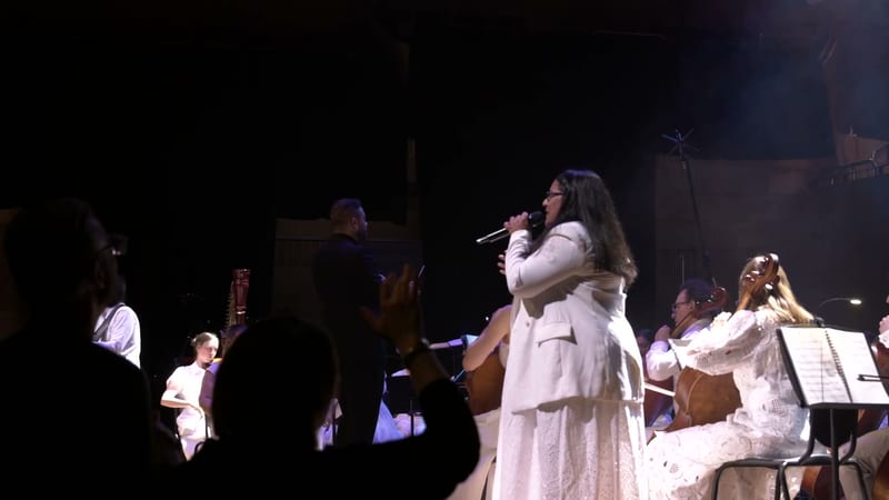 A soloist in white performs with microphone while accompanied by string players and orchestra members on stage.