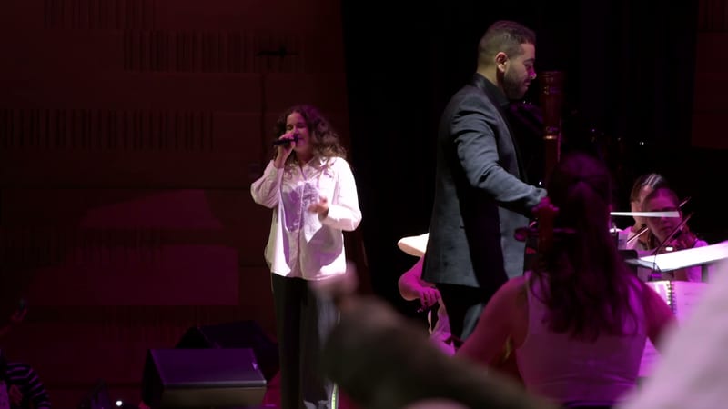 A female soloist sings at the microphone while a male performer gestures expressively on stage, with orchestra members visible in the background.