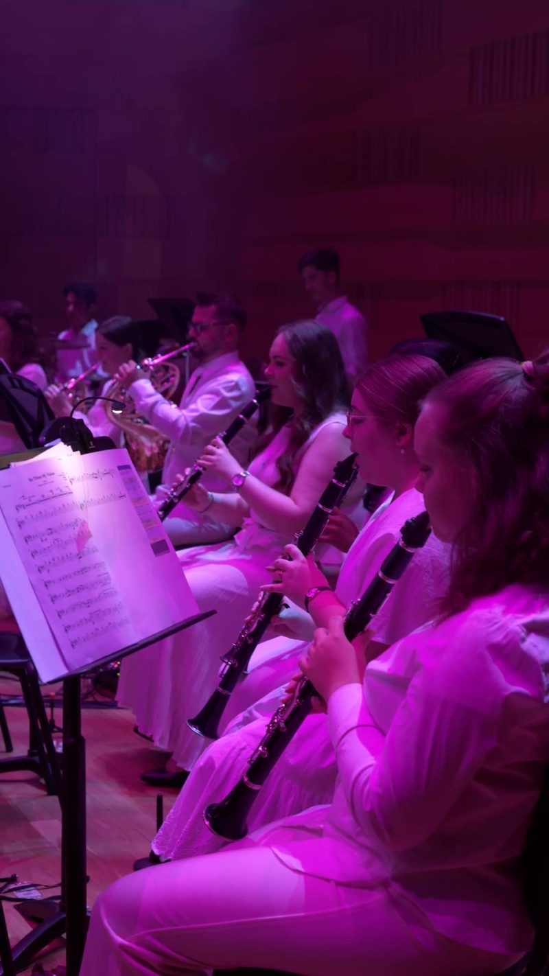 Woodwind section of young musicians performing under magenta stage lighting with sheet music stands.