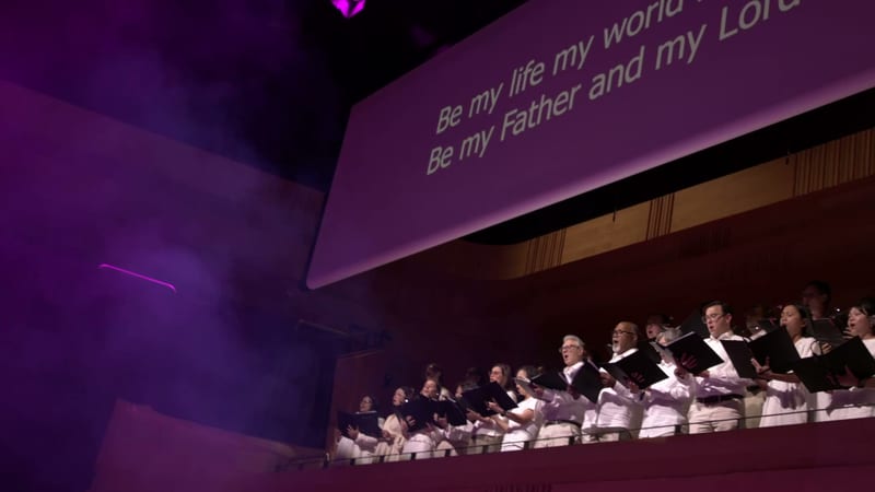 A choir performs in formal white and black attire beneath a purple-lit screen displaying spiritual text.