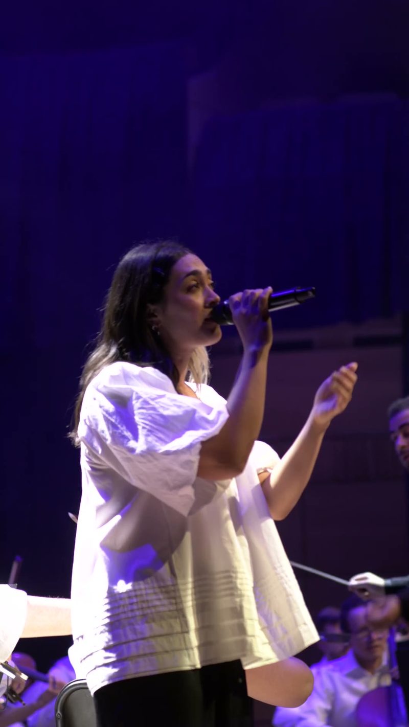 A female soloist performs at the microphone on stage, wearing a white shirt and illuminated by purple stage lighting.