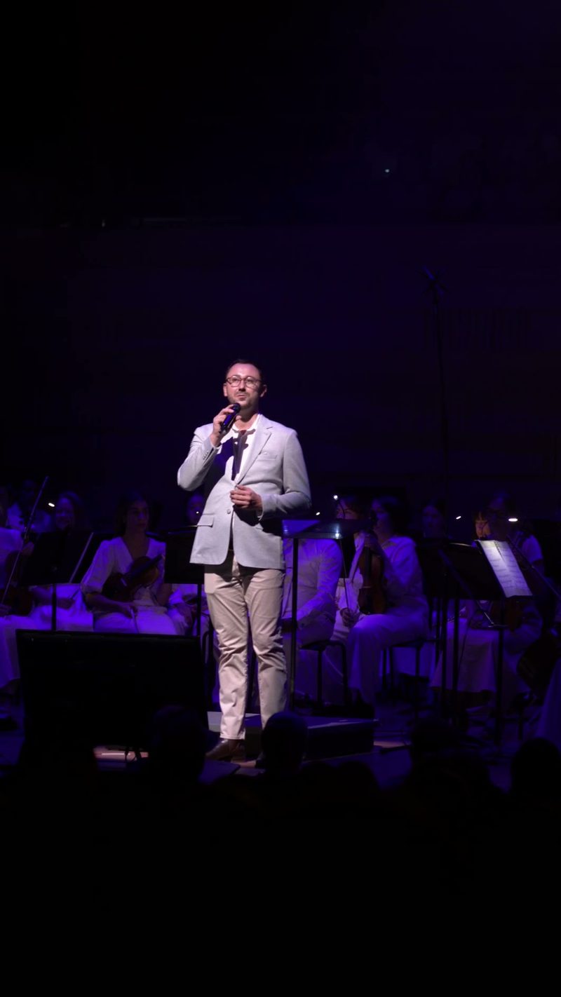 A male soloist in a light gray blazer speaks into a microphone while performing with an orchestra under purple stage lighting.